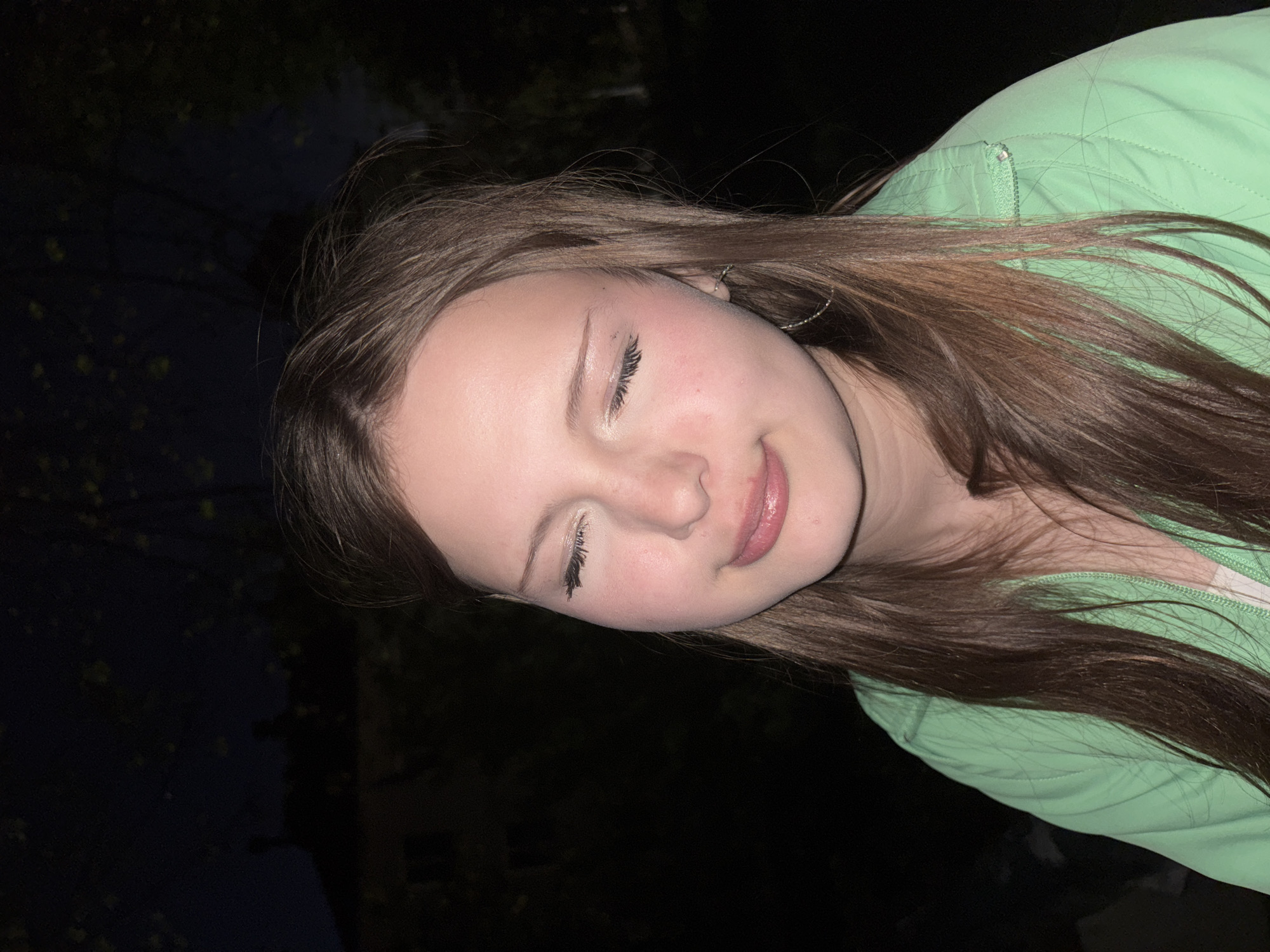 The image shows a young woman with long brown hair. She is wearing a light green jacket and has a silver hoop earring in her left ear. Her eyes are closed and she appears to be smiling slightly. The background is dark, with some blurry shapes suggesting trees or buildings. There is no information to determine a location in the city of Фоки, Russia.