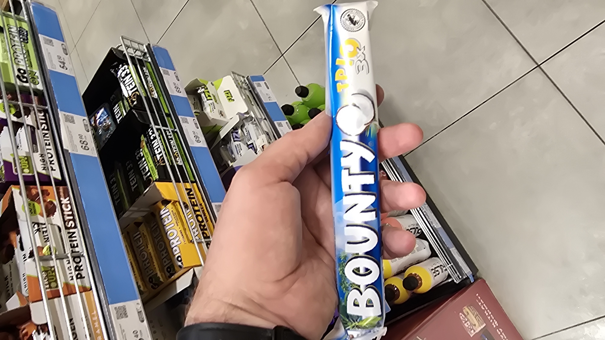 A person's hand is holding a Bounty Triple Chocolate bar in front of a shelf of protein bars. The person's hand is in the foreground, and the Bounty bar is angled slightly upwards. The Bounty bar has a blue wrapper with white lettering that says "Bounty" and "Triple Chocolate". There is also an image of a coconut on the wrapper. In the background, there are shelves filled with various protein bars and other snacks. The shelves are made of metal, and the products are arranged in boxes and individual packages. The overall scene appears to be in a supermarket or convenience store. The floor is tiled with light-colored tiles.