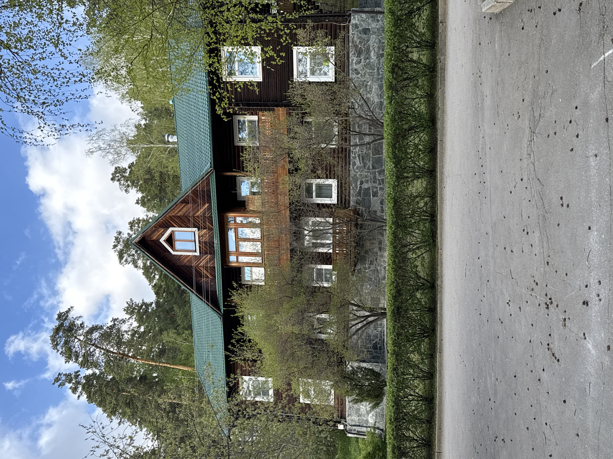 The image depicts a multi-story wooden building with a green tiled roof, likely located in a wooded area. The building has numerous windows of varying sizes and a triangular gable facing the viewer. Part of the facade appears to be constructed with stone. In the foreground, there is a paved road adjacent to a row of neatly trimmed bushes. Trees surround the building and are visible in the background along with a partially visible blue sky with clouds. The location is Zolotoi Plyazh, Russia, though there are no specific identifiers in the image itself. There are no people visible in the image.