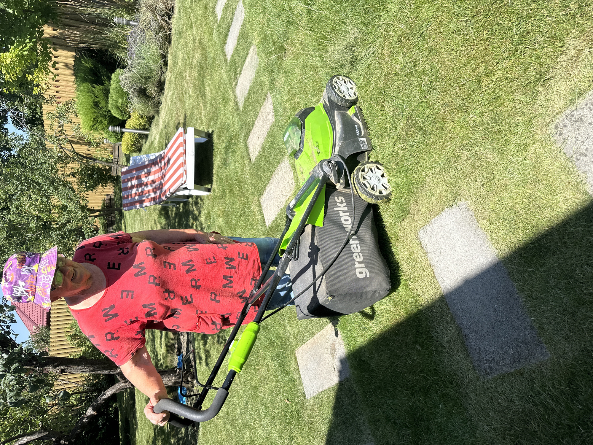 In Armavir, Russia, on a sunny day, an elderly woman with glasses and a purple bucket hat stands in a well-maintained backyard, preparing to mow the lawn with a green and black electric lawnmower. She is wearing a red t-shirt with black lettering and blue jeans. The lawnmower, a "greenworks" model, has a collection bag attached. The backyard features a neatly mowed green lawn, a stone pathway, a wooden fence, trees, bushes, and a red and white striped lounge chair.