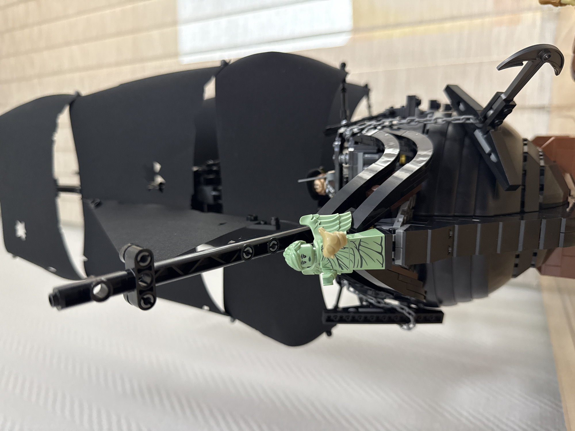 The image depicts a Lego model of the Silent Mary pirate ship from the movie "Pirates of the Caribbean: Dead Men Tell No Tales." The ship is primarily black, with tattered black sails. A light green Lego minifigure of the Statue of Liberty is attached to the bowsprit. The background appears to be a blurry indoor setting, likely near a window with blinds. The location cannot be determined as Krasnoyarsk, Russia from the provided information.