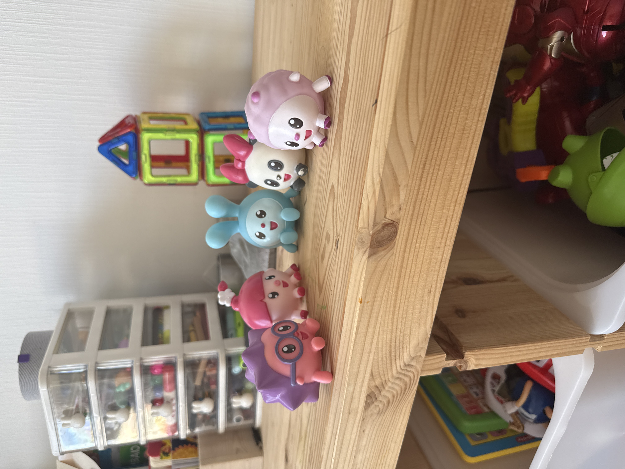 This is an indoor, eye-level medium shot of a wooden table with toys on it, against a white wall. The subjects of the photo are five small, colorful plastic toys arranged on the table in the foreground. From left to right, they are: a purple, spiky creature wearing glasses; a pink, bell-shaped creature; a light blue bunny; a white and black panda with a pink bow; and a light pink, round creature. In the background, to the right of the toys, is a stack of magnetic building blocks. To the left of the toys, a multi-drawer plastic storage unit is visible, filled with various small items. The wooden table has a visible wood grain pattern and is slightly worn. Below the table, the edge of a white storage bin is visible, containing colorful toys. The overall environment appears to be a child's play area. The location is not specified in the image.