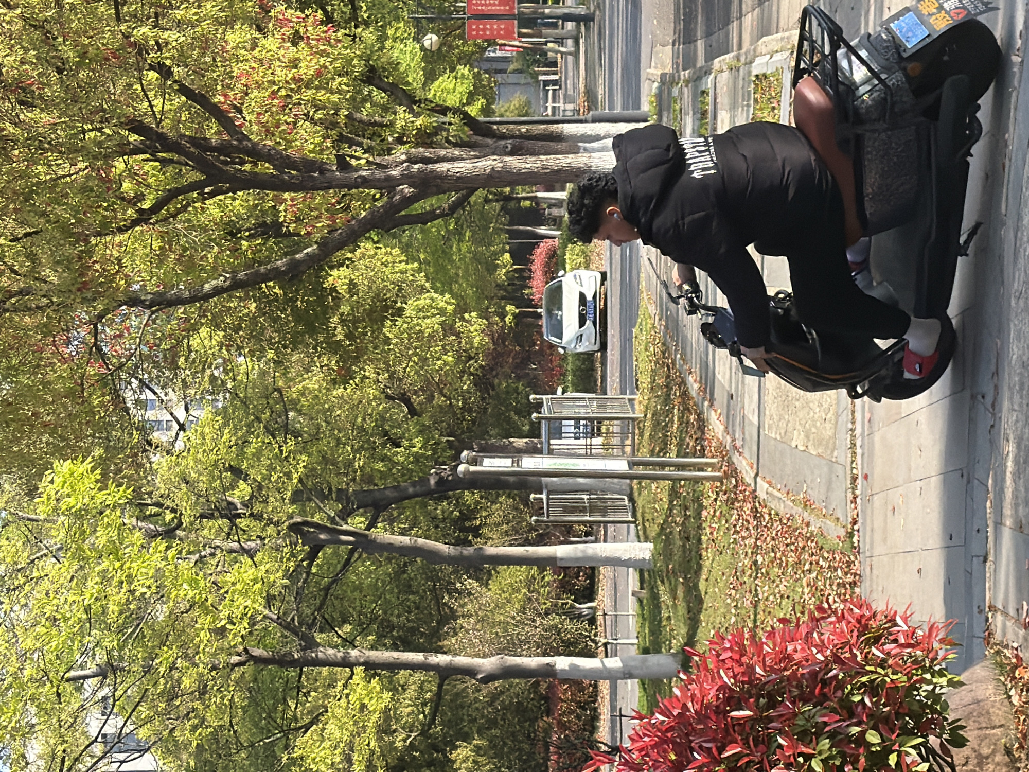 A man wearing a black jacket, black pants, and red shoes is seen on a scooter on a sidewalk. The scene is bordered by trees with green leaves and some red bushes. A white car is visible in the background on the road, and a sign with Chinese characters hangs above the road further away. The location is Jiangning District, China, based on the sign.