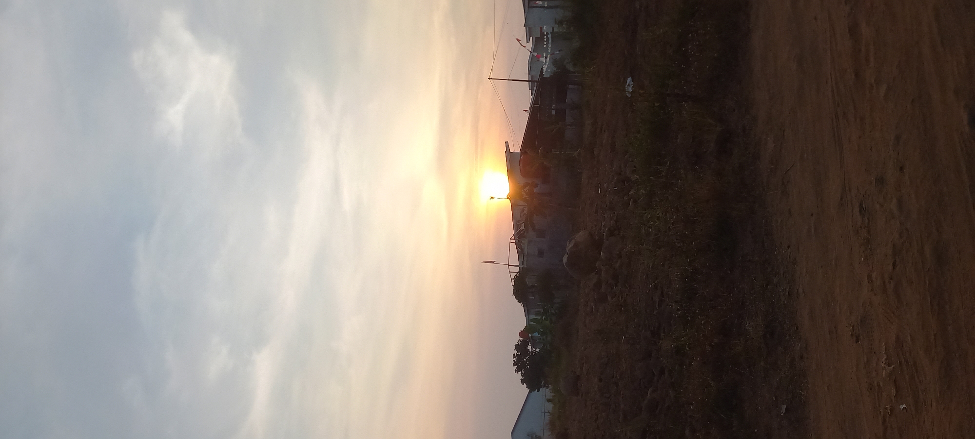 The image depicts an outdoor scene at sunset in Loa Lepu, Indonesia. The sky is a gradient of pale blue and orange, with soft clouds, indicating the sun is low on the horizon and casting a warm glow. In the middle ground, several buildings with pitched roofs are visible, one appearing under construction. A small red and white flag is mounted on one of the structures. The foreground consists of uneven, brown earth with sparse vegetation, suggesting undeveloped land or a construction site. There are large rocks scattered on the ground. The overall atmosphere is tranquil and rural.