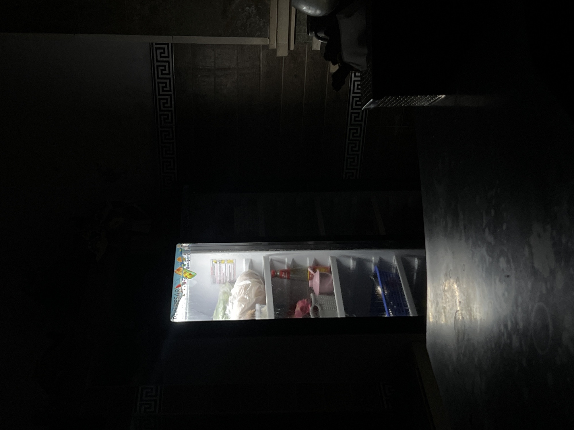 The image shows the interior of a dimly lit space in Canggu, Indonesia. The primary focus is an open refrigerator displaying various food items and containers. Beyond the refrigerator, there's a partially visible countertop or surface. In the background, some steps can be seen and a person or mannequin is standing or leaning. The overall environment is dark, with the refrigerator's light being the main source of illumination. Greek key pattern borders can be seen on the walls.