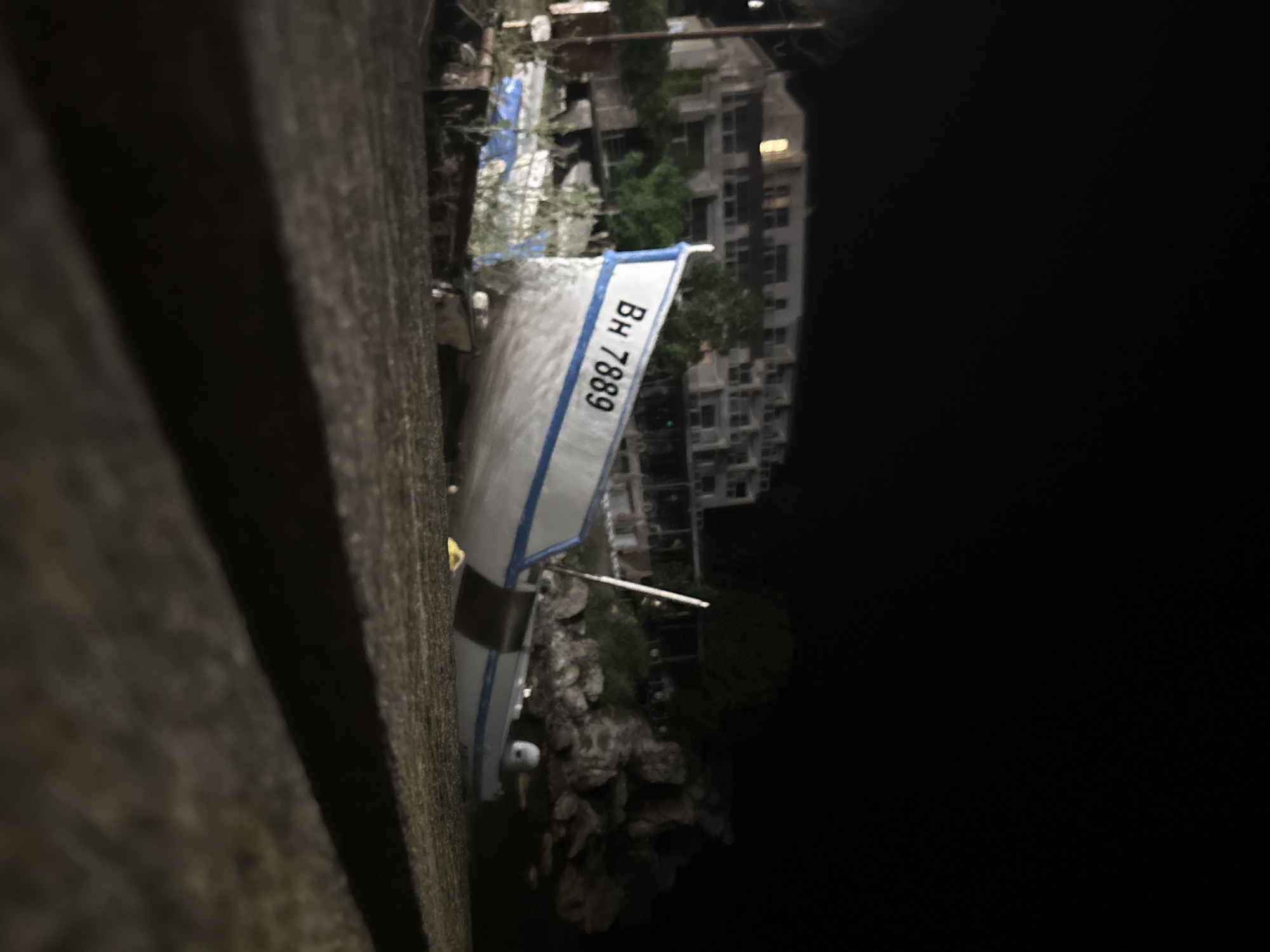 At night in Tyulenovo, Bulgaria, a white fishing boat with blue trim is beached on the shore. The boat has the registration number "BH 7889" painted on its side. Behind the boat, a multi-story building with illuminated windows is visible, suggesting it is a hotel or residential complex. The surrounding environment includes rocks, vegetation, and dark sky. The foreground is a blurred, dark surface. There are no people visible in the image.