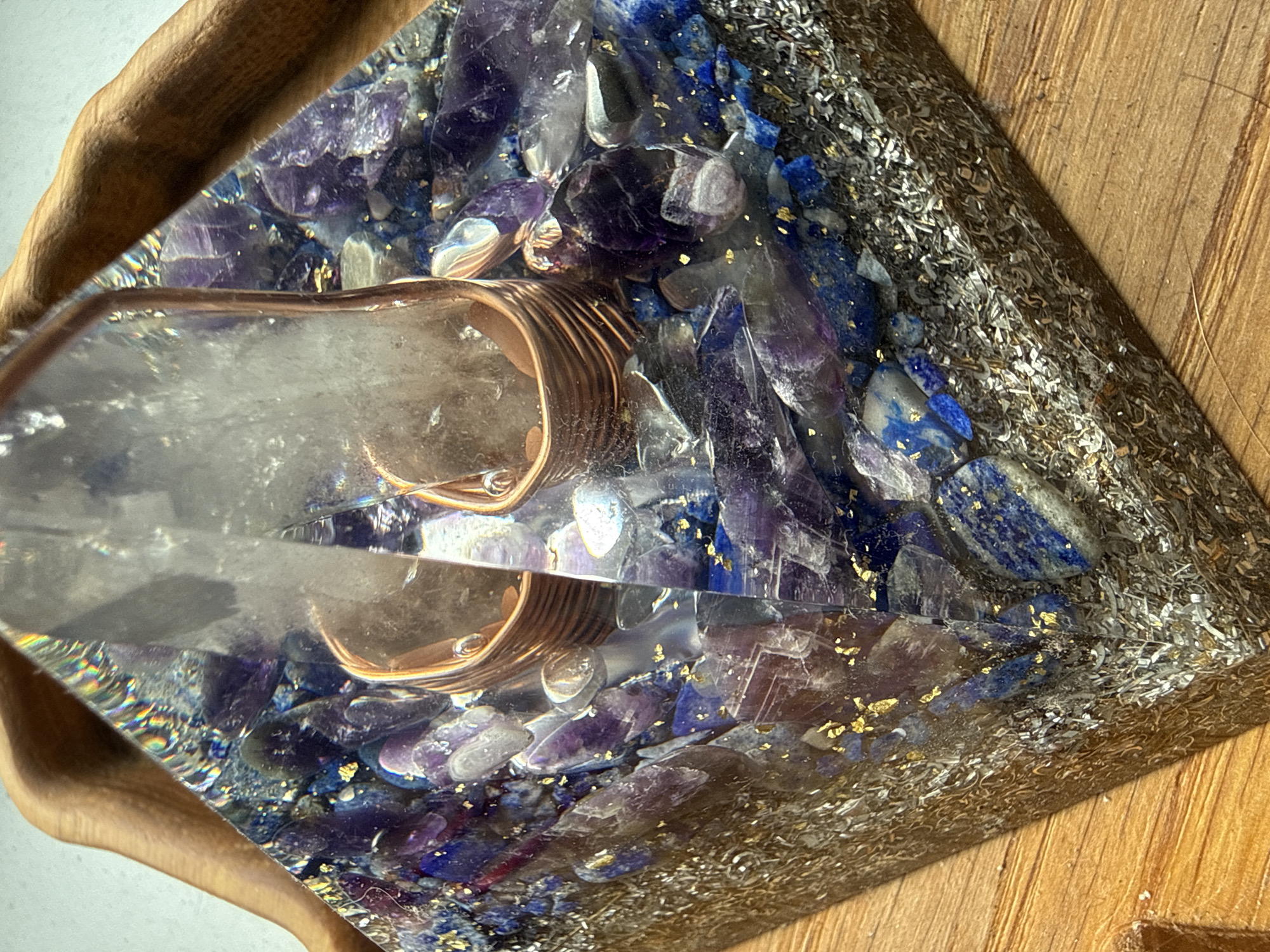 An orgonite pyramid with amethyst and lapis lazuli crystals is displayed on a wooden surface. The pyramid is partially obscured by a curved piece of wood.
