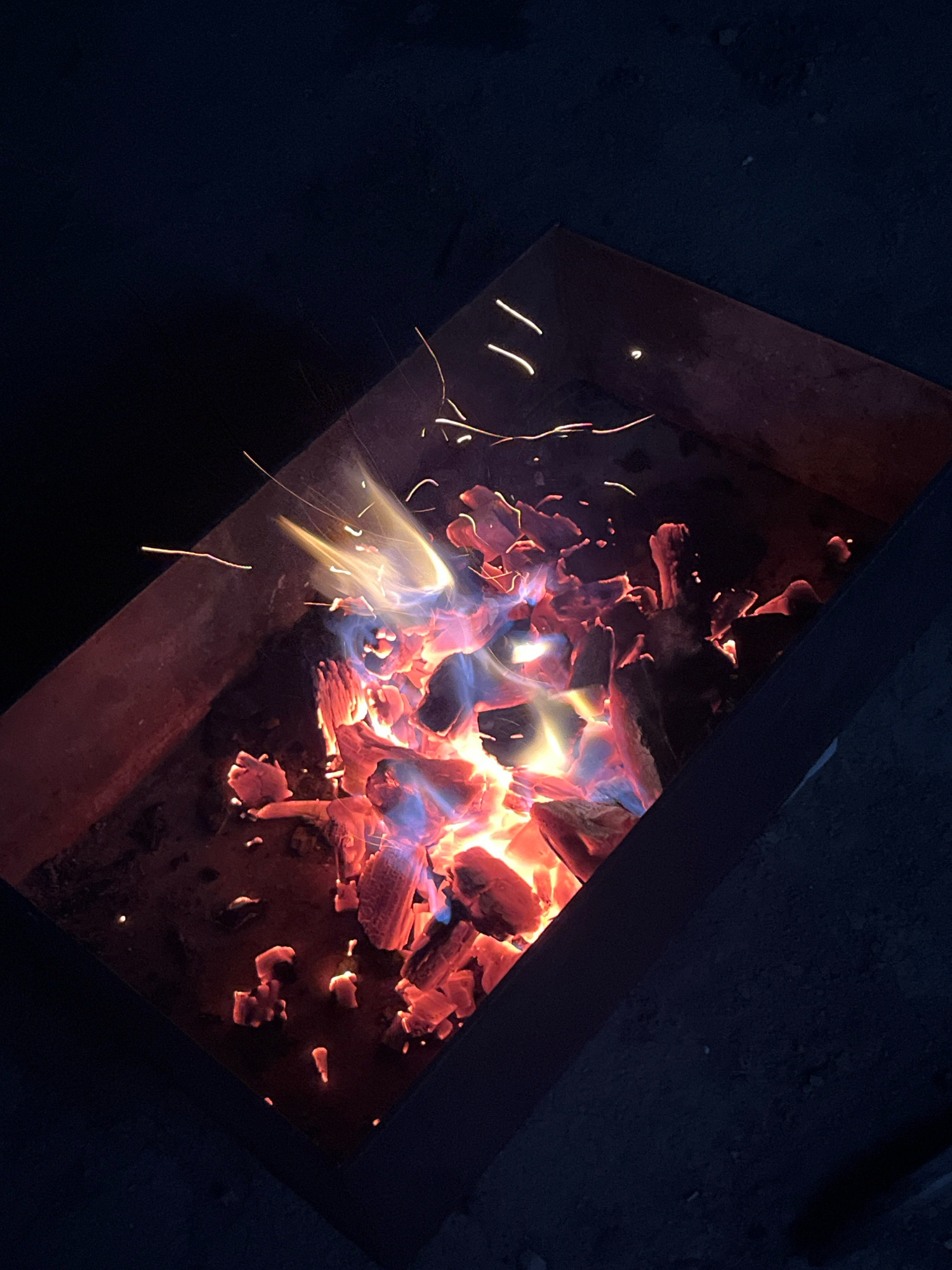 A close-up, high-angle view shows a metal fire pit filled with glowing red and blue embers and burning wood. Sparks are visible flying from the fire. The fire pit is set against a dark, out-of-focus background. The scene appears to be outdoors at night.
