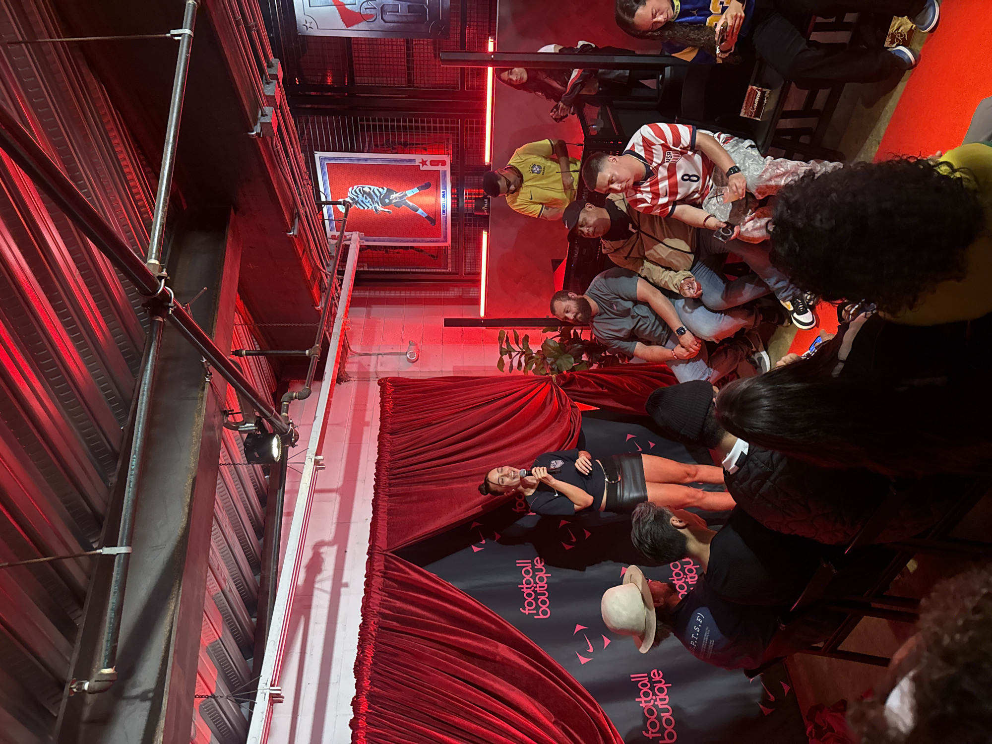 The image depicts an indoor event, likely in Los Angeles, United States of America. A woman stands on a small stage speaking into a microphone. The stage is adorned with red curtains and features a backdrop with the words "football boutique" printed on it.
Seated in front of the stage is an audience of approximately 10 people. The attendees display varying styles of dress.
The indoor space has a raw, industrial aesthetic, with exposed metal beams, scaffolding, and red lighting accentuating the environment. A framed poster hangs on the wall behind the audience.