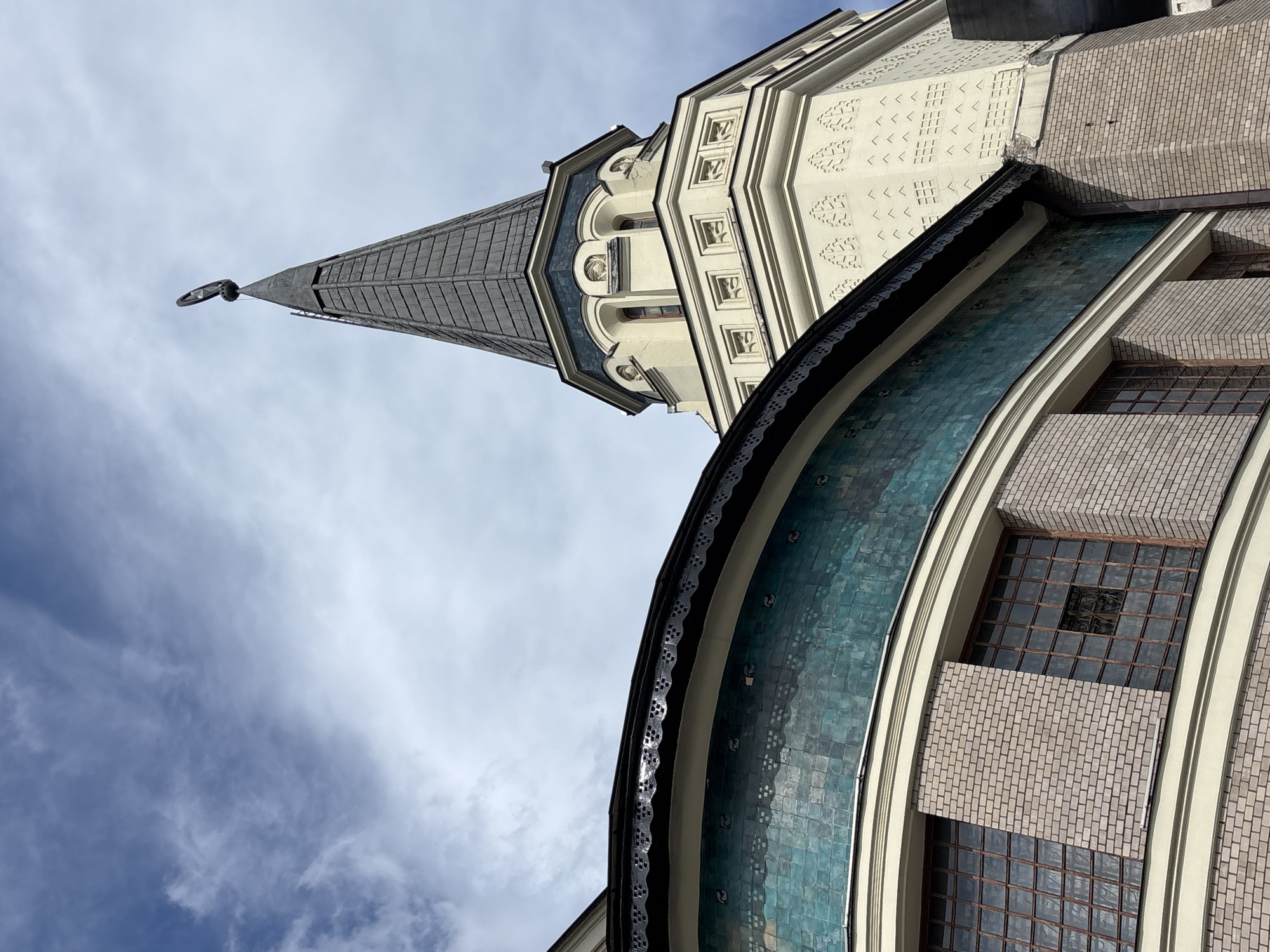 The image depicts an upward view of architectural details of a building in Moscow, Russia, against a partly cloudy sky. A tall, pointed spire, likely a tower, is centrally located. The spire features a gray, shingled roof culminating in a decorative element at its peak. Adjacent to the spire is a cream-colored structure with patterned details. In the foreground is a rounded, tiled section of the building, exhibiting a turquoise hue and a dark, decorative edge. The windows are rectangular and appear to have a grid pattern. The architecture appears to be detailed and potentially of historical significance.
