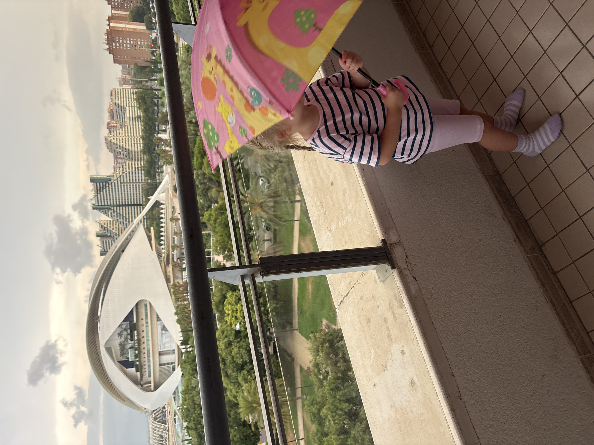 A young girl stands on a balcony holding a pink umbrella with giraffes on it. She is wearing a striped t-shirt and light purple shorts, along with striped socks. The balcony overlooks the City of Arts and Sciences in Valencia, Spain, with its distinctive architecture visible in the background. The sky is cloudy.