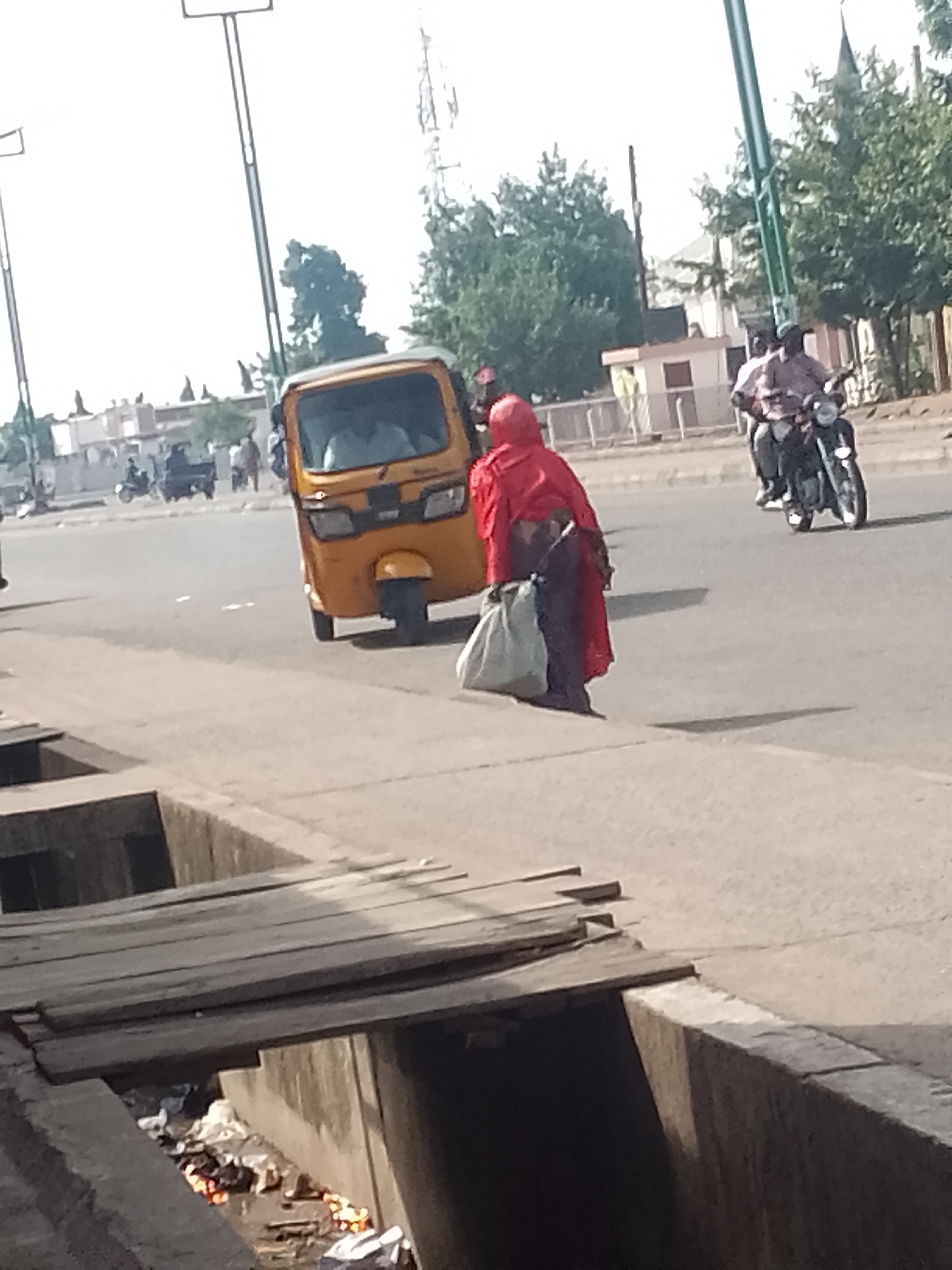 This image displays a street scene in Pantame, Nigeria, observed during daylight hours. In the immediate foreground, a concrete drainage ditch traverses the frame, containing accumulated debris and visible glowing orange/red embers, indicative of smoldering refuse. Several wooden planks are laid across portions of this ditch, forming an improvised crossing.
Adjacent to the ditch, a paved sidewalk or shoulder extends. A pedestrian, clad in a red outer garment, is depicted walking away from the camera, moving rightward along the sidewalk. This individual carries two bags, one light-colored and one dark-colored.
On the asphalt road surface in the midground, a yellow three-wheeled auto-rickshaw is positioned at an angle, suggesting either recent movement or a stop. Further along the road, a motorcycle carrying two occupants is visible, proceeding in the same general direction. Shadows cast on the road surface indicate the presence of direct sunlight.
The background consists of various low-rise buildings and scattered trees. Multiple street light poles are present, and a prominent metal telecommunication tower is visible in the upper right quadrant of the image, set against a bright, hazy sky.