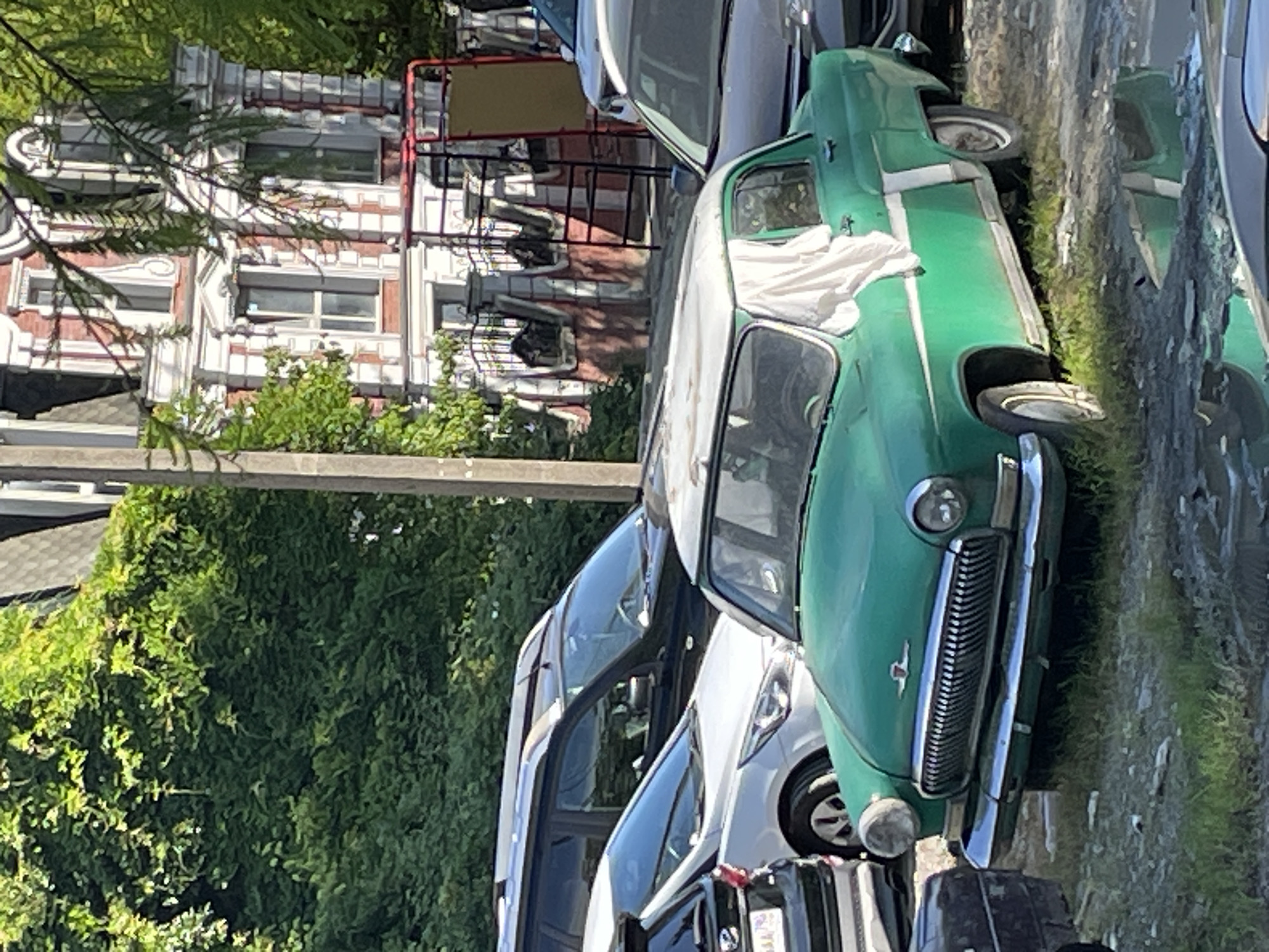 A vintage green car with a white roof is parked on a gravel lot. The car has a chrome grille and bumper, and a white sheet is draped over the passenger side window. In the background, there is a large building with red and white striped accents and a red metal structure. Lush green trees and foliage are visible throughout the scene. The car is reflective in a puddle on the ground. The scene appears to be in Sochi, Russia.