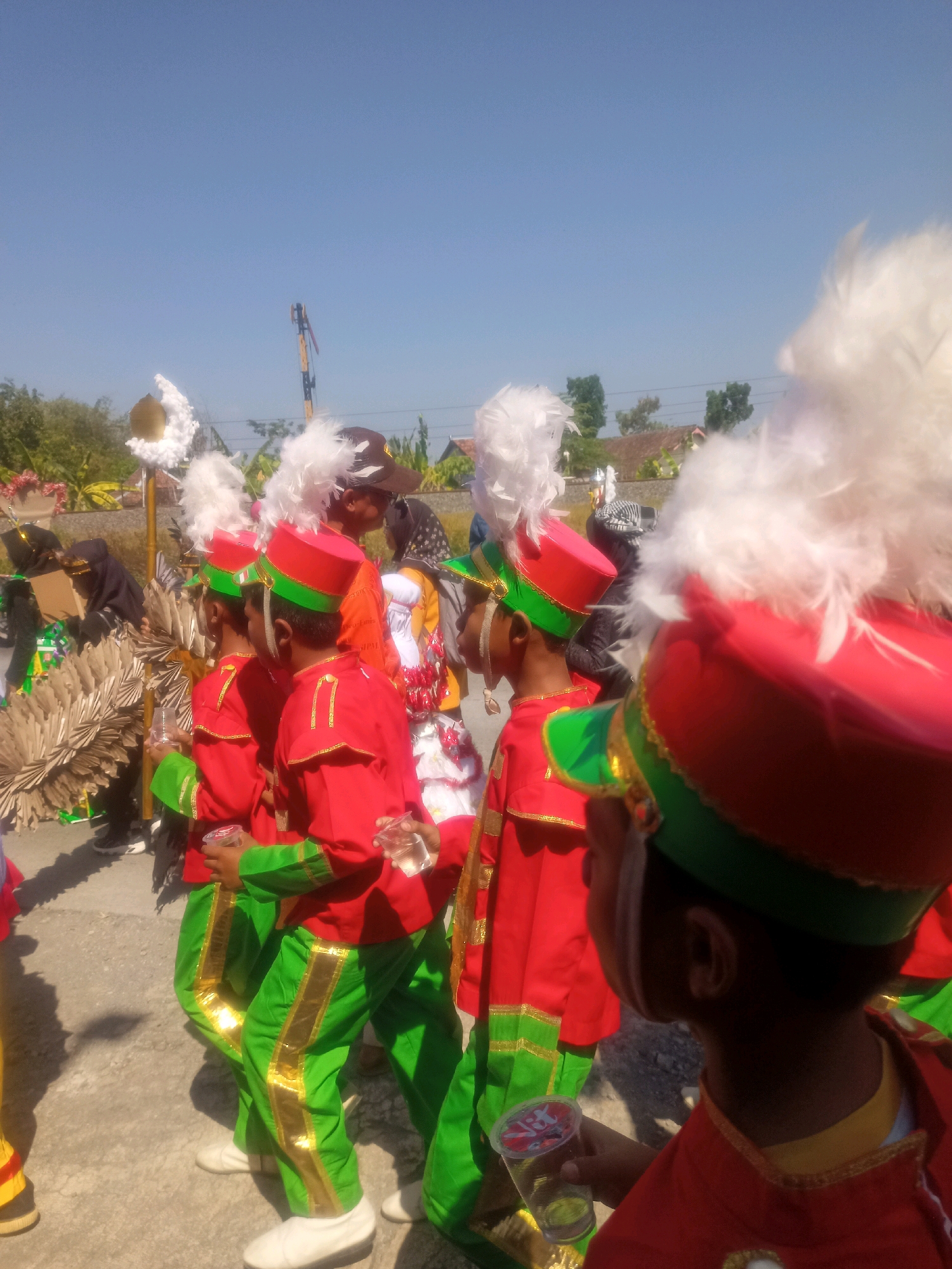 In Sumberlawang, Indonesia, on a sunny day, a group of children in marching band uniforms with red jackets, green pants with gold stripes, and red hats adorned with white feathers are seen participating in a parade or event. Some of the children are holding clear plastic cups. In the background, there are trees, buildings, and a utility pole. The ground appears to be paved.