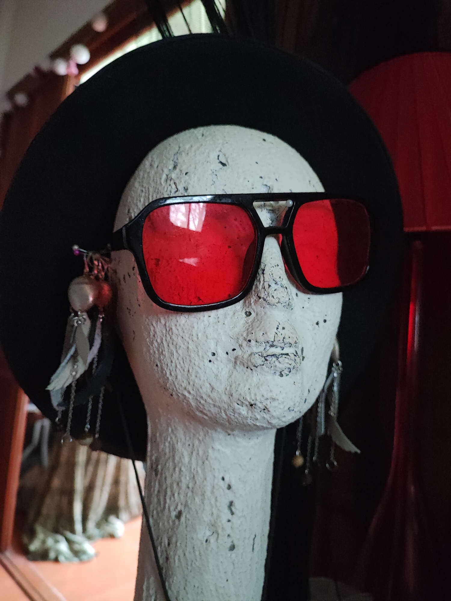 A mannequin head wearing a black hat and red-tinted sunglasses is displayed against a dark background. The mannequin has a textured white surface with dark speckles. Attached to the hat above the mannequin's ears are two decorative earrings. The earring on the left appears to be composed of a round, light-colored bead, silver chain links, and light-colored feathers. The earring on the right has a similar beaded and chain structure, along with a light-colored feather. The background is mostly dark, with hints of red fabric or a lamp on the right side of the image. To the left, a blurred background shows a window with translucent curtains and decorative white spheres, suggesting an indoor setting, potentially a shop or display area. No specific location in Ko Pha-ngan District, Thailand, can be determined from the visual information.