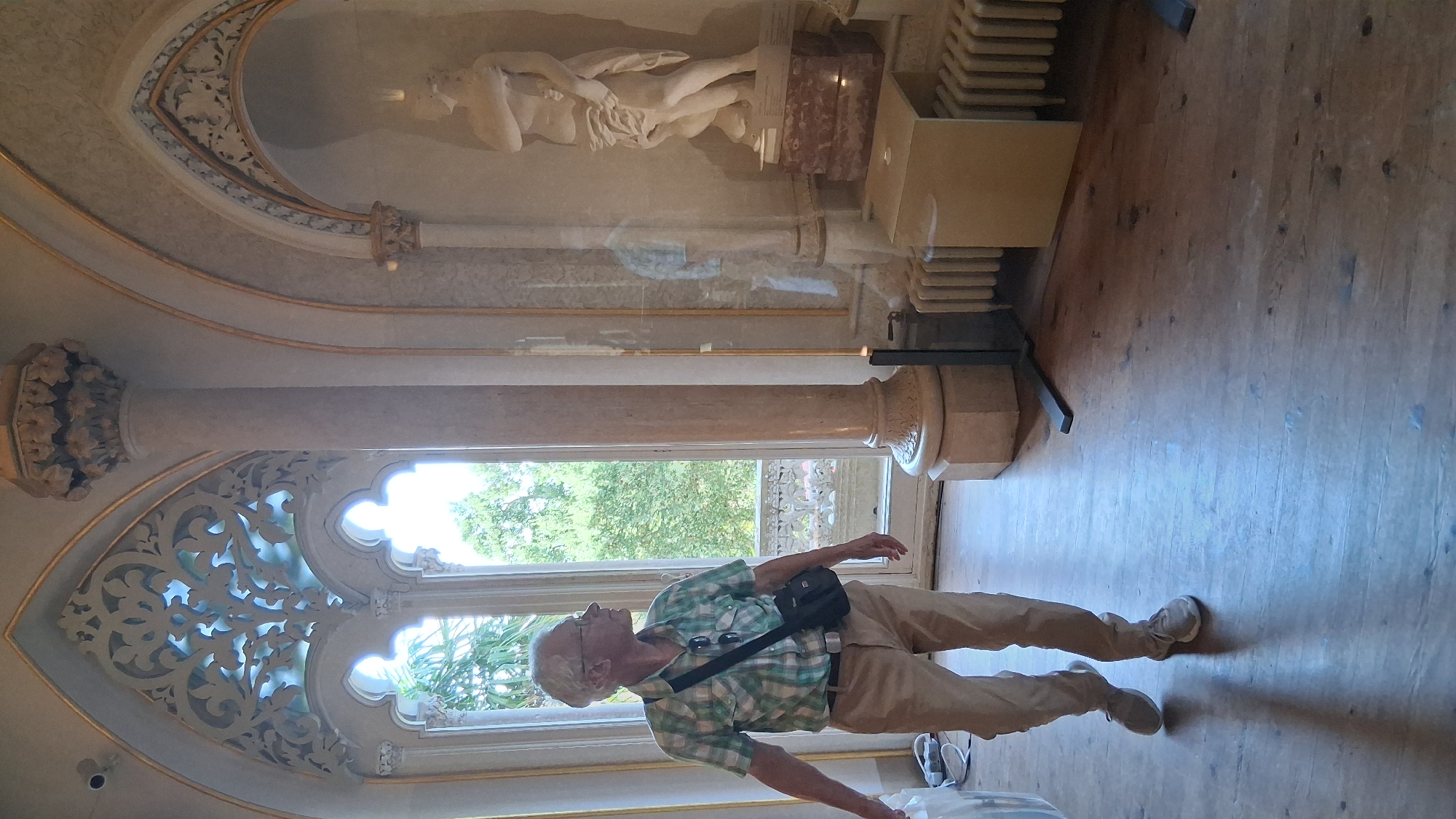 An elderly man with gray hair and glasses, wearing a plaid shirt and light-colored pants, walks through a room with ornate architectural details. He has a dark shoulder bag across his chest. The room features a large arched window with intricate carvings and a view of lush green trees. A tall, fluted column is visible on the right, and an arched alcove contains a white sculpture of a draped figure. The floor is made of weathered wooden planks. The scene appears to be inside a historic building, possibly part of Pena Palace in Sintra, Portugal, given the distinctive architecture and decorative elements.