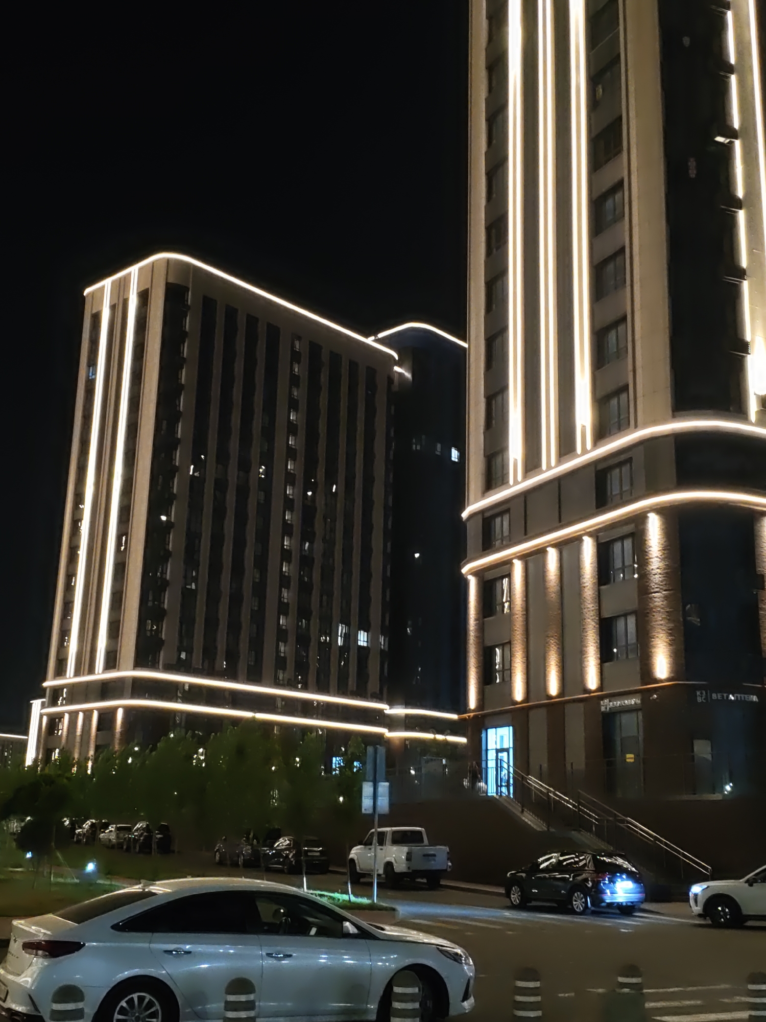 This is a nighttime street-level photograph taken in Rostov-on-Don, Russia. The scene features two modern, illuminated high-rise buildings with prominent vertical and horizontal lighting accents. In the foreground, a silver sedan is parked. Several other vehicles, including a white pickup truck and a dark SUV, are visible on the road and in parking areas. Green trees line the street, and the ground is paved. The lighting suggests an urban environment with businesses or residences in the illuminated buildings.