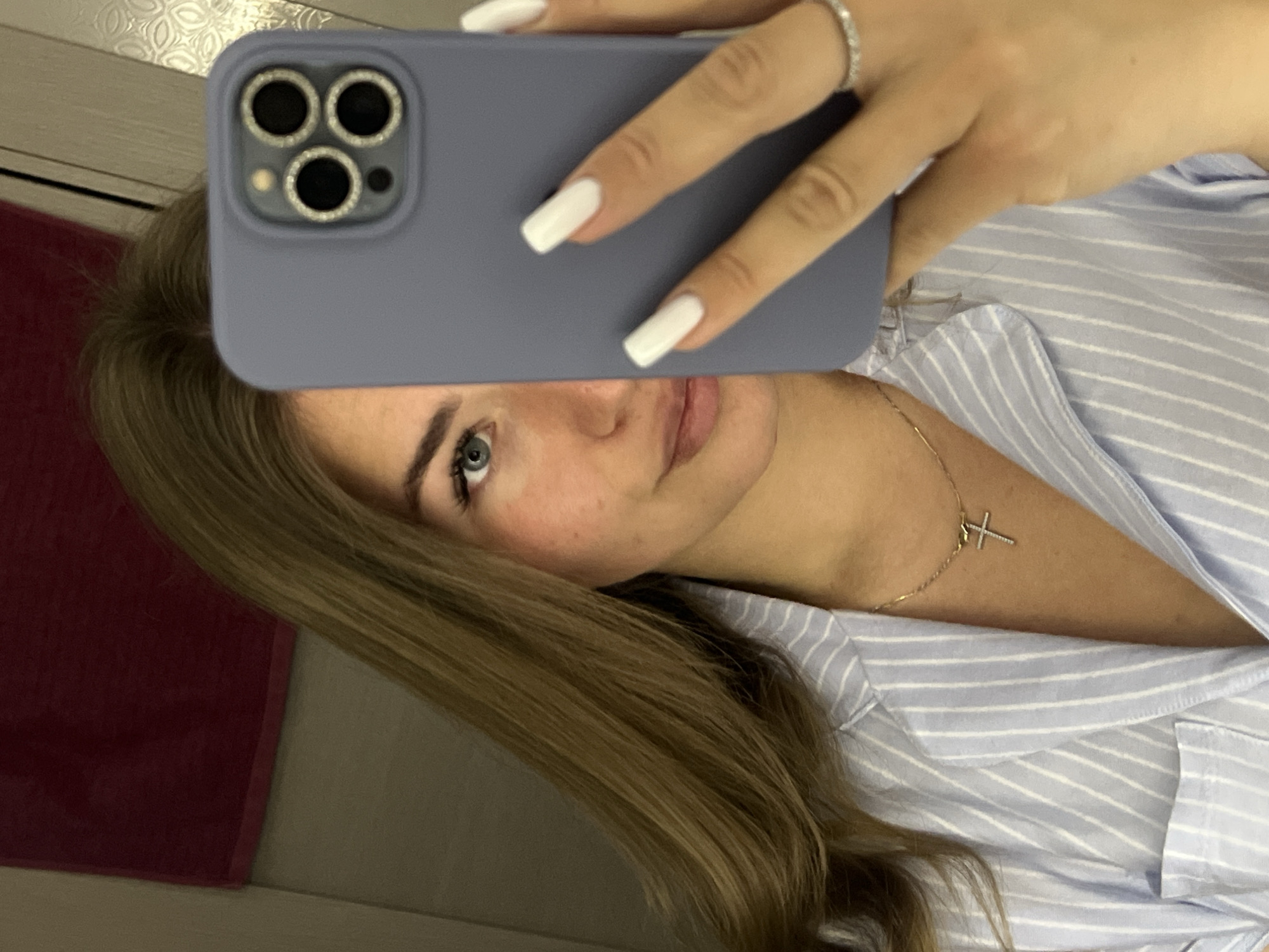 An indoor mirror selfie captures a young woman, partially obscured by a smartphone, in what appears to be a private residence. The subject has long, light brown hair, fair skin with some freckles, and a visible left eye of blue-green color. Her lips are slightly parted. She is wearing a light blue shirt with white vertical stripes and a gold chain necklace with a small, silver-colored cross pendant adorned with tiny stones. Her right hand, positioned in the foreground, holds a smartphone vertically, covering the right half of her face. The phone is encased in a pale blue/lilac protective cover, featuring a prominent triple-lens camera module on the rear, each lens surrounded by a sparkling, possibly jeweled, ring. Her right hand displays long, square-tipped white fingernails and a thin silver-colored ring with small stones on her ring finger. The background indicates an interior space: a dark red towel hangs on a light-colored wall to the upper left, while a reflective surface with a decorative patterned detail is visible on the upper right, possibly a mirror frame or furniture. The specific location within the city of Budva, Montenegro, cannot be determined from the visual evidence in the image.
