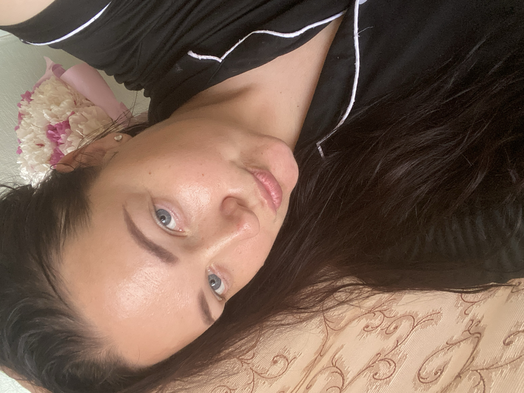A woman with fair skin, light blue eyes, and dark hair is reclining on a surface with a beige floral pattern. She is wearing a black pajama top with white piping. A bouquet of flowers is visible near her head. The image does not provide enough information to determine if she is in Sochi, Russia.