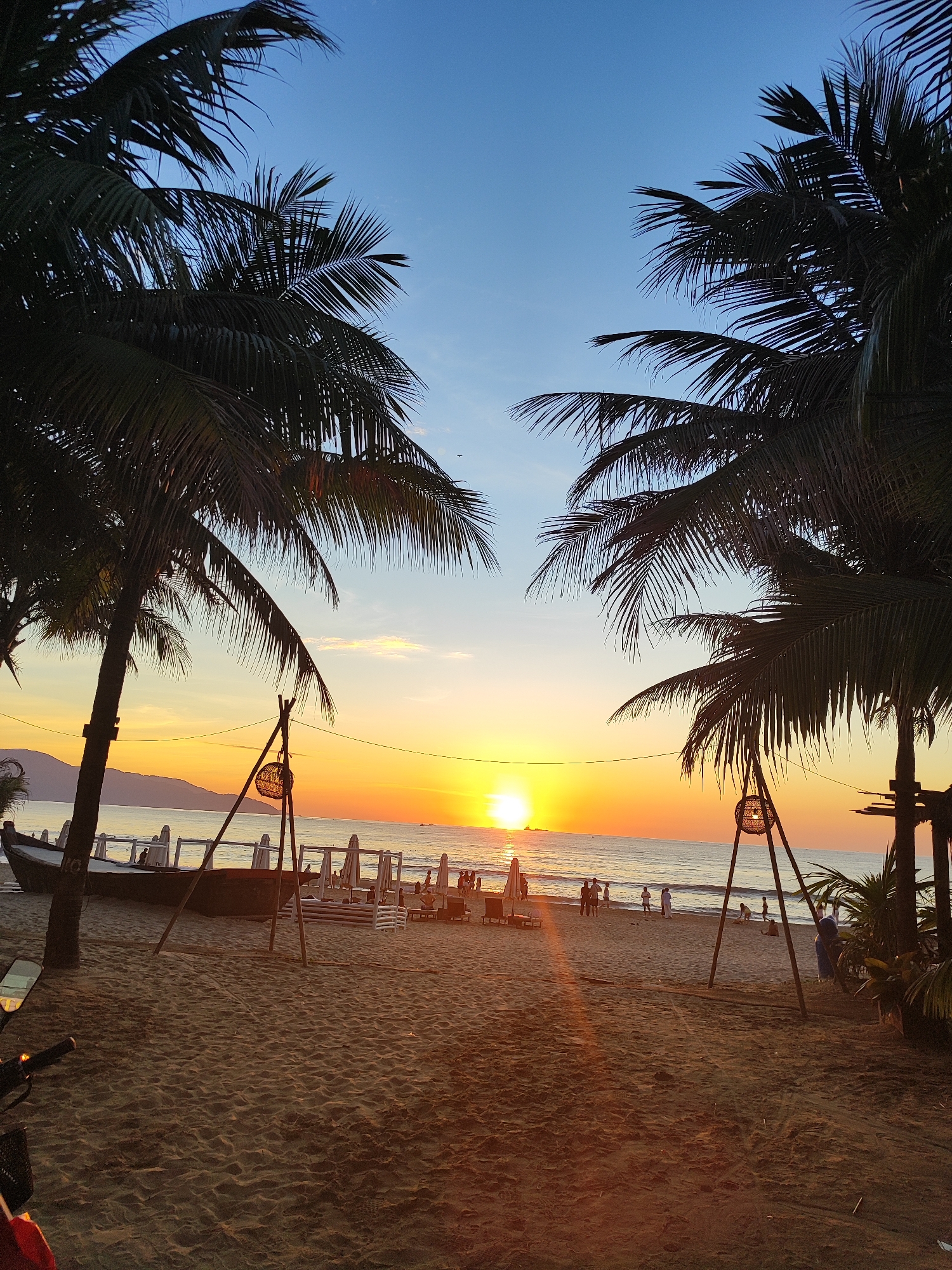 A picturesque sunset scene unfolds on a sandy beach in Đà Nẵng, Vietnam, as viewed through the silhouettes of two palm trees. The sky transitions from a deep blue at the top to a vibrant orange and yellow gradient near the horizon, where the sun is prominently displayed as a bright orb. The calm sea reflects the warm hues of the sunset, and a faint line of hills or mountains can be seen in the distance.
In the foreground, the sandy beach is dotted with various objects. To the left, a portion of a motorcycle is visible, hinting at recent arrival or departure. A long, boat-like structure, possibly a decorative element or a repurposed vessel, lies on the sand. Several white beach umbrellas are erected, and lounge chairs are set up, suggesting a resort or public beach area. Two wooden tripods, each with a woven lantern hanging from them, stand on the sand, adding to the ambiance.
Further back, a group of people are scattered along the shoreline, some walking, some standing, and a few appearing to interact. They are small figures, silhouetted against the bright backdrop of the sunset. The overall atmosphere is serene and tranquil, capturing a moment of natural beauty and leisurely beach life.
