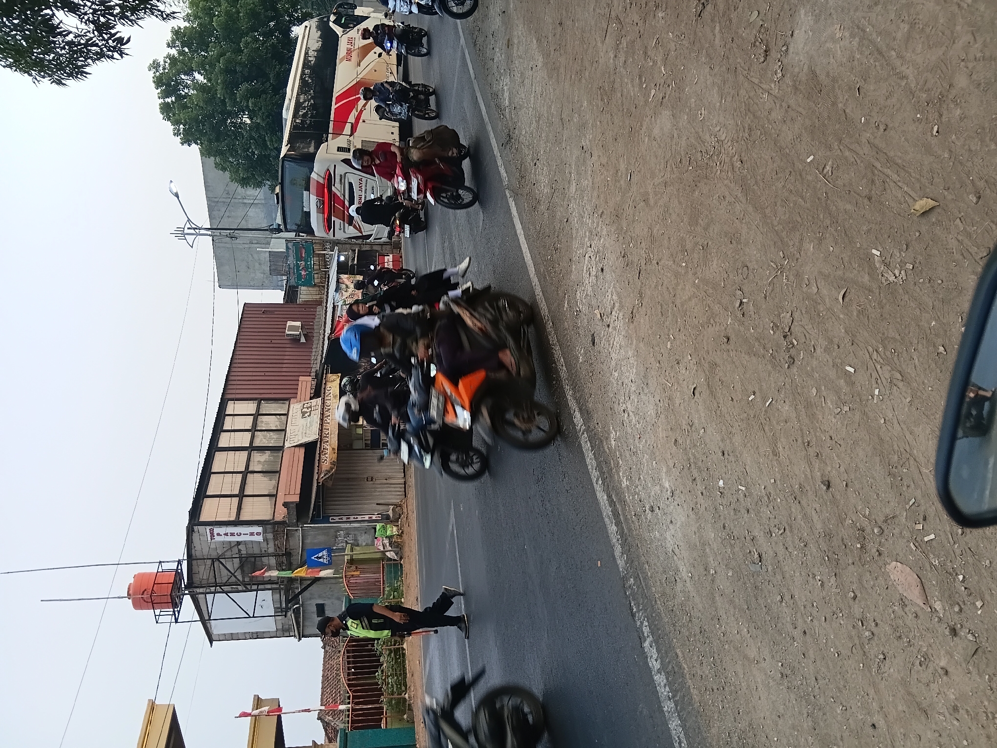 This is a street-level, medium-shot photograph taken in Bekasi, Indonesia, featuring a busy road with numerous motorbikes and a large bus. The scene appears to be an urban or semi-urban environment, with buildings and infrastructure visible on the left side of the road.
In the foreground, the road surface is visible, with tire tracks and some debris. The right edge of a black mirror is partially visible in the bottom right corner, suggesting the photo might have been taken from a vehicle.
On the left side of the road, a man in a dark uniform with a bright green vest is walking across the street, holding a long stick or pole. He appears to be directing traffic or performing some form of security duty. Behind him, a fence and buildings are visible, including a structure with a large orange water tank on top. There are also flags and signs on the buildings.
The road itself is filled with traffic. Multiple motorbikes are in motion, with some riders blurred due to their speed. The motorbikes vary in color, with a prominent orange one in the center foreground. Several riders are wearing helmets.
In the background, a large white and red bus with the text "MURNI JAYA" is prominently displayed. The bus is also in motion. Trees and other vegetation are visible further in the background, along with more buildings and what appears to be a street light. The sky is overcast and light. The overall impression is one of a typical day on a busy Indonesian road.