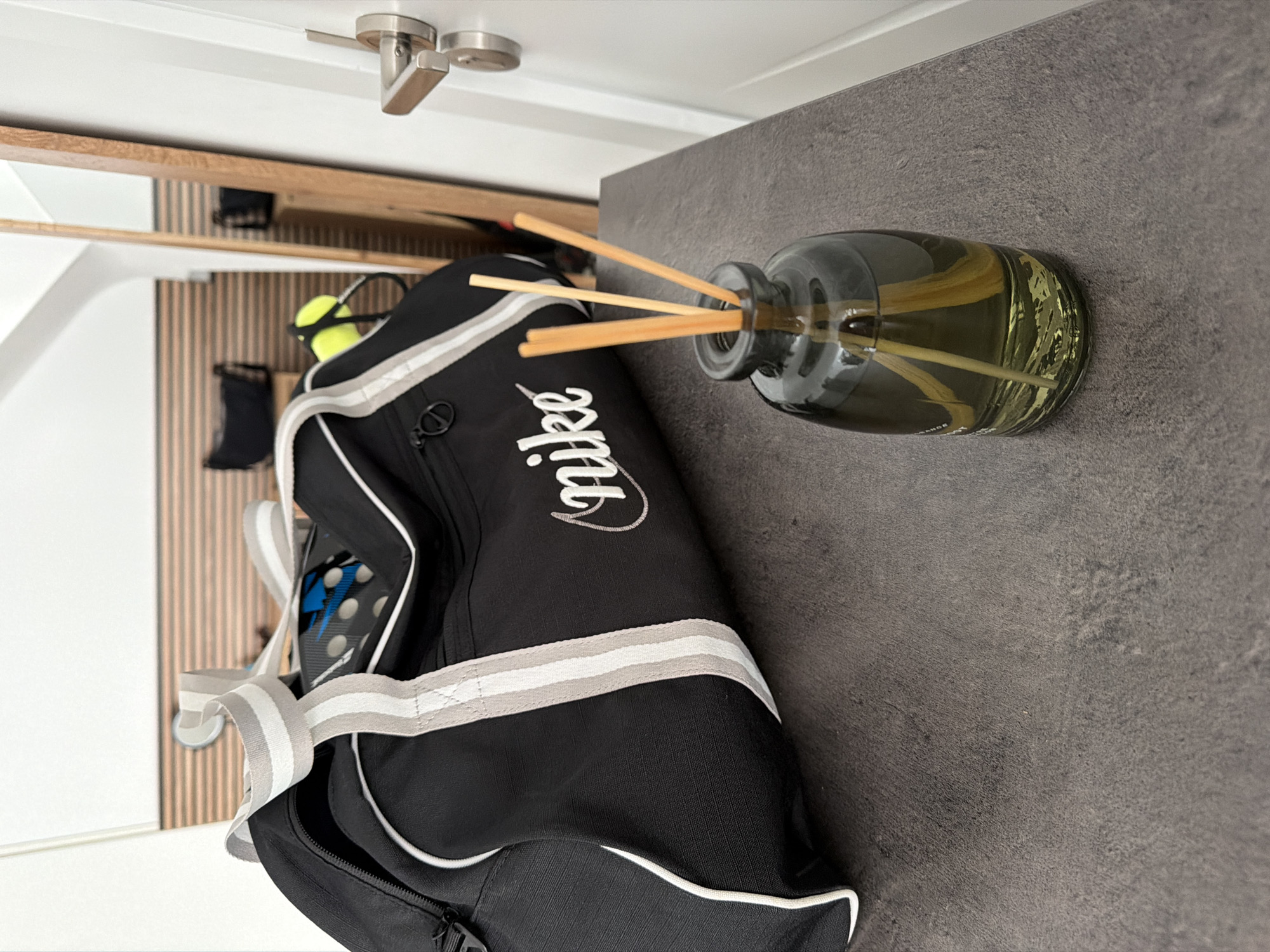 In Finestrat, Spain, a black Nike duffel bag with white and gray stripes sits on a dark gray textured countertop. To the right of the bag is a small glass bottle containing a pale green liquid and several thin wooden reeds, likely a reed diffuser. In the background, a tall mirror with a wooden frame reflects a room with wooden-paneled walls and a black handbag on a surface. A silver doorknob is visible on a white door to the right of the frame. The lighting suggests an indoor setting, possibly a hallway or entryway, with natural light coming from the right. There are no people or animals visible in the scene.