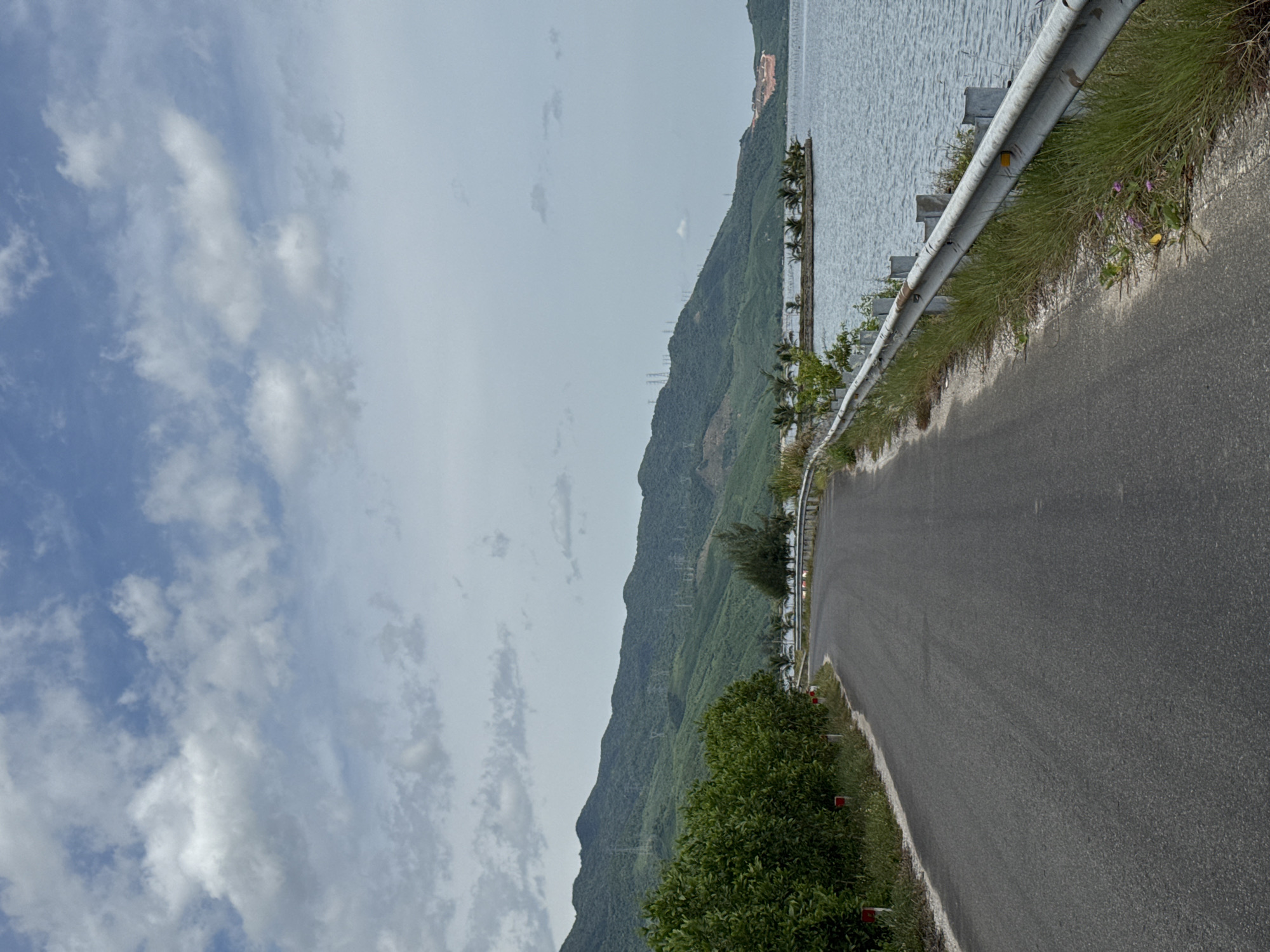 The image depicts a road curving alongside a body of water, with lush green hills and mountains in the background. The road is paved and appears to be an asphalt surface. Along the right side of the road, there is a metal guardrail with grass and some small pink flowers growing beneath it. The body of water is calm and appears to be a lake or bay, with a small island dotted with palm trees visible in the distance. The mountains are densely covered in vegetation, and transmission towers can be seen on their slopes. The sky is partly cloudy with a mix of blue sky and white clouds. The scene is likely located in the outskirts of Huế, Vietnam, given the characteristic landscape of the region which often features coastal roads, mountains, and bodies of water. There are no people or animals visible in the image. The overall atmosphere is serene and natural.