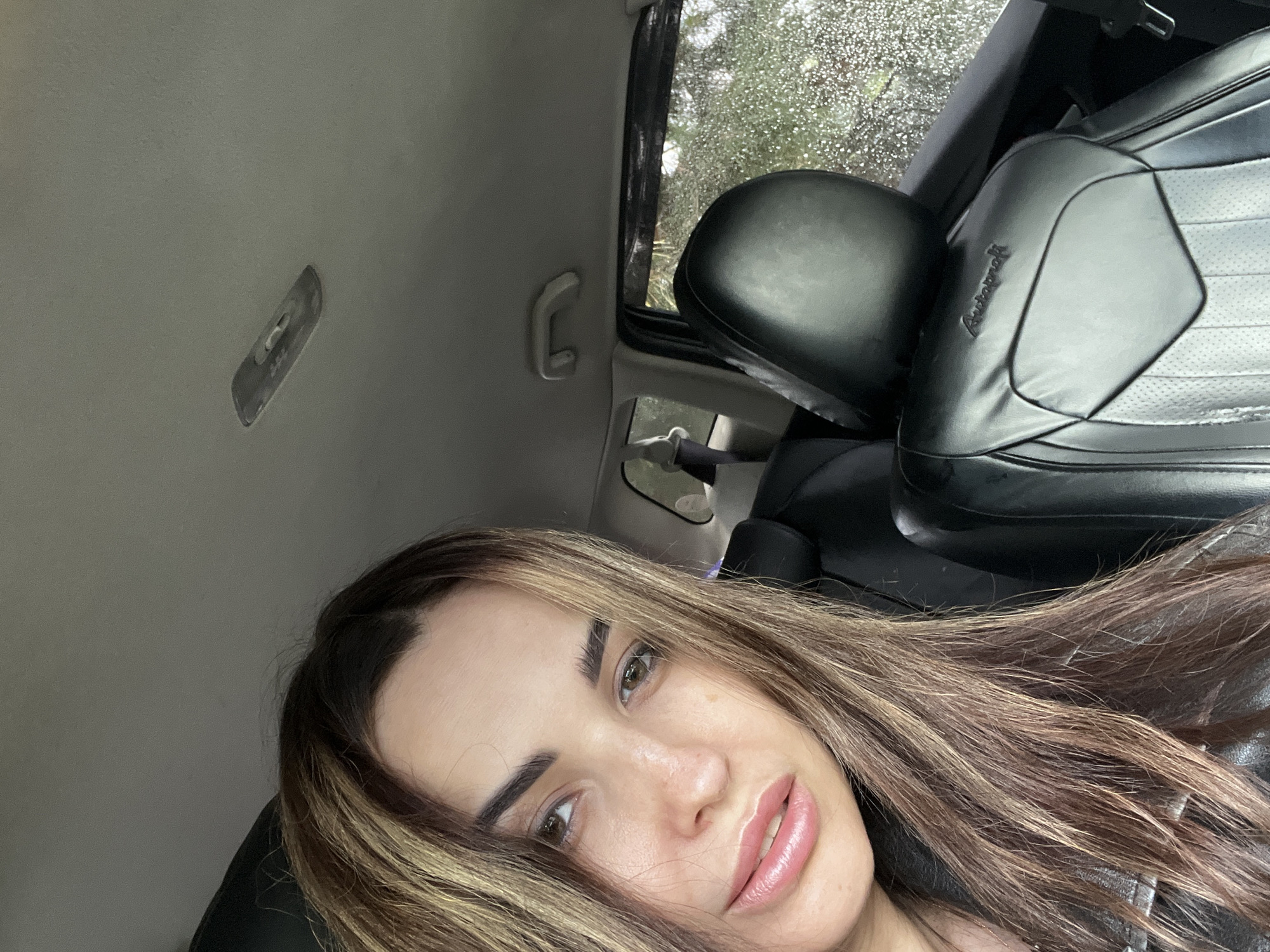A woman takes a close-up selfie from inside a car. Her face, with brown eyes, well-defined eyebrows, and light pink lips, dominates the lower right portion of the frame. She has long, wavy brown hair with caramel highlights, which flows across her shoulder.
The setting is a car interior. The light grey headliner and an overhead light/control panel are visible above her. To her left, a white grab handle is attached to the car's pillar. Through the car window, numerous raindrops are visible, indicating rainy or recently rainy weather, with blurred greenery or trees outside. A black car seat, featuring both smooth and perforated sections, is prominently displayed on the right side of the image. The word "Autocraft" is embossed on the seat. A black headrest and part of a seatbelt are also visible. The woman's expression is neutral to slightly contemplative. The scene appears to be taking place during the daytime.