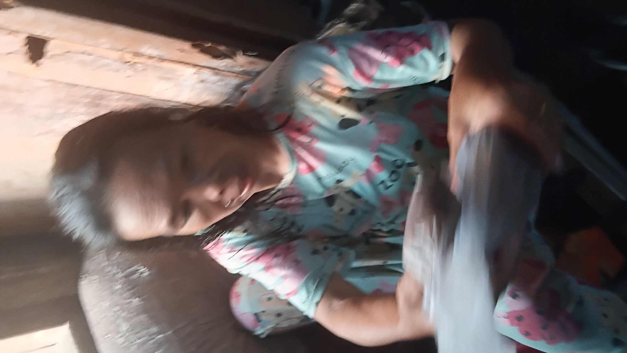 A blurry, medium shot captures a woman sitting and holding a clear plastic bag filled with a dark substance, possibly food or produce. She is wearing a light blue pajama set with a pink and black cartoon animal pattern, featuring what appears to be pandas and puppies. The background is dimly lit and appears to be an interior space with textured walls and possibly furniture. The overall image quality is low, with significant motion blur, making it difficult to discern specific details of the environment or the woman's facial features. The scene is not definitively identifiable as being in Prabumulih, Indonesia.