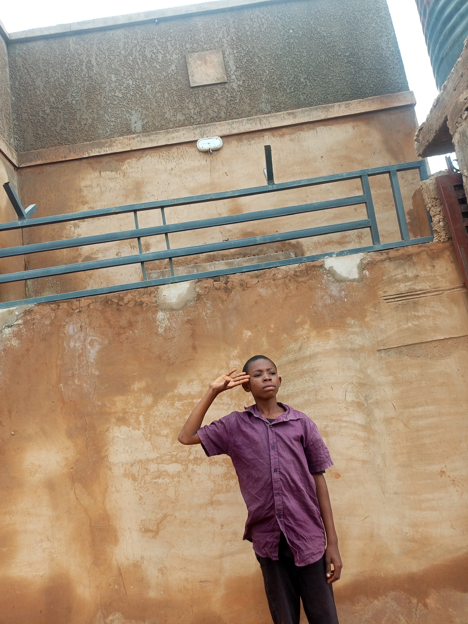 A young male, appearing to be of African descent, stands in front of a weathered, ochre-colored wall. He is wearing a purple, short-sleeved button-down shirt and dark trousers. His right hand is raised to his forehead in a salute-like gesture. Above and behind him is a grey metal railing fixed to a textured grey structure. The overall scene suggests an outdoor environment, potentially in a residential or urban setting, with elements of building construction and wear visible. The location is identified as Tempure, Nigeria.