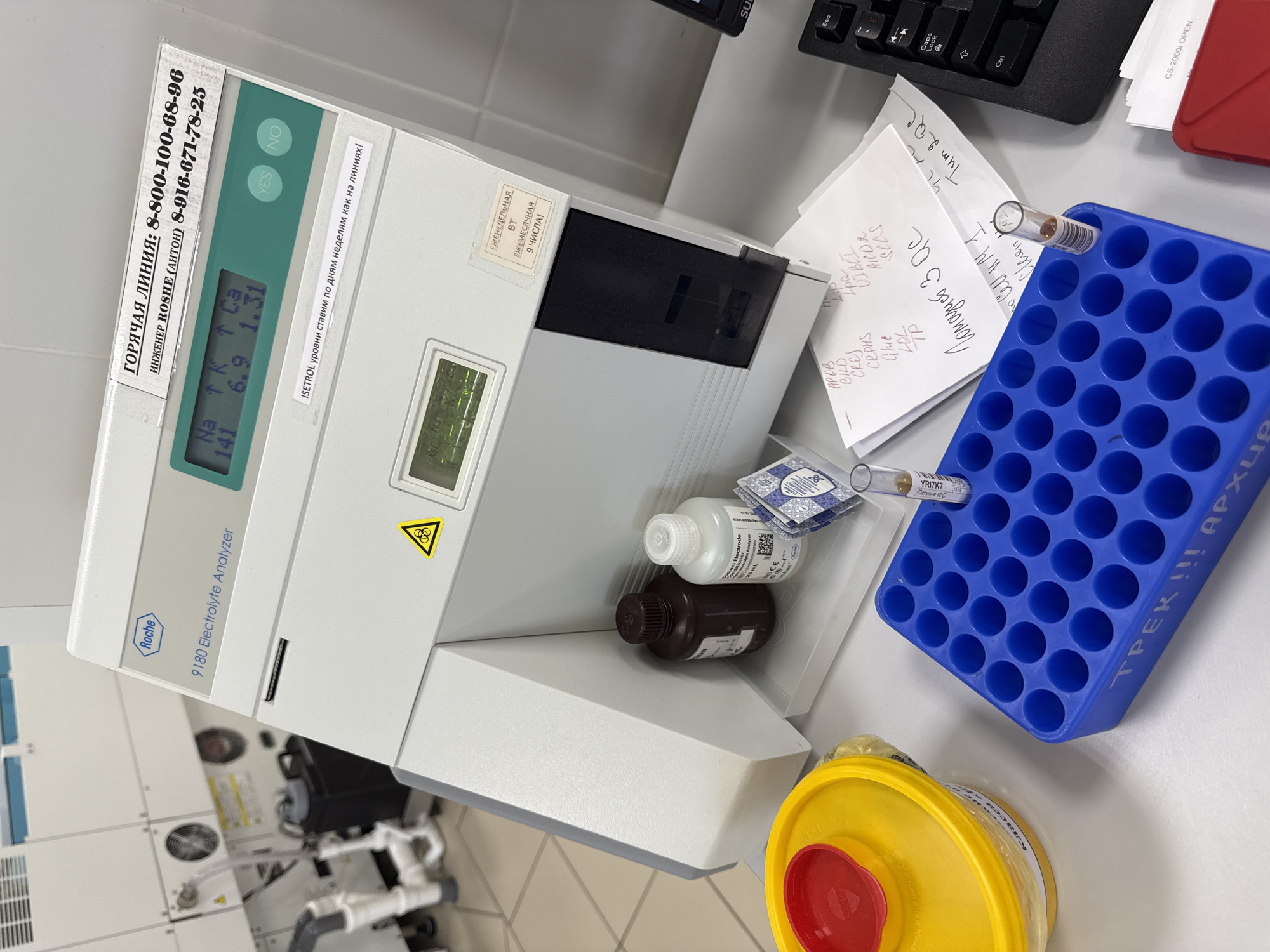 The image depicts a laboratory setting in Moscow, Russia, showcasing a Roche 9180 Electrolyte Analyzer. A blue test tube rack holds several blood samples, with handwritten notes alongside. Waste disposal containers are present near the analyzer. The analyzer's display shows electrolyte levels for Na, K, and Ca. Bottles of solutions are visible on top of the analyzer. A computer keyboard is in the background.