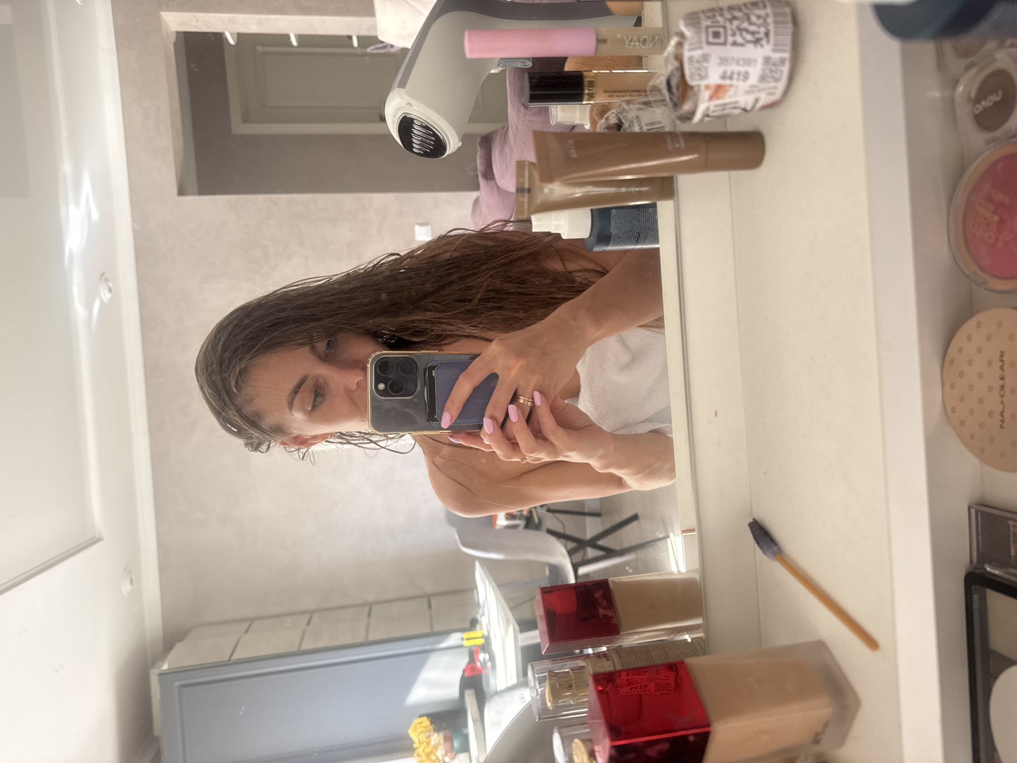 A woman is seen taking a mirror selfie in what appears to be an apartment in Sochi, Russia. Her long, dark brown hair is wet, and she is wrapped in a white towel around her chest. She holds a smartphone with a prominent multi-lens camera array (suggesting a recent iPhone model) in both hands, positioning it to capture her reflection. Her fingernails are painted light pink, and she wears two thin, gold-colored rings on her left ring finger. Her gaze is directed downward towards the phone screen, not directly at the camera.
The immediate foreground of the image shows a white vanity or countertop. On it are two foundation bottles (one with a red square top, the other a clear pump bottle containing a light beige liquid), a slender makeup brush with a dark tip, and a crumpled piece of paper with a QR code and numerical labels ("257629", "4419"). A white and grey handheld garment steamer is also visible on the right side of the counter. Reflected behind these items are additional beauty products including brown tubes, spray bottles, and a pink cylindrical item. Below the counter, partially visible, are several makeup compacts, including one marked "NAJ OLEARI".
The reflection in the mirror reveals the apartment's interior. To the left, a grey cabinet, a sunlit window, and a bouquet of yellow flowers are visible. A light-colored table and two modern white chairs are present in the background. The wall features a textured, light beige finish, with a doorway leading to another room and a light-colored door. Further back, a soft, purple-mauve sofa or armchair is visible. The ceiling is white, equipped with recessed lighting and a decorative rectangular panel. A white light switch is mounted on the wall.