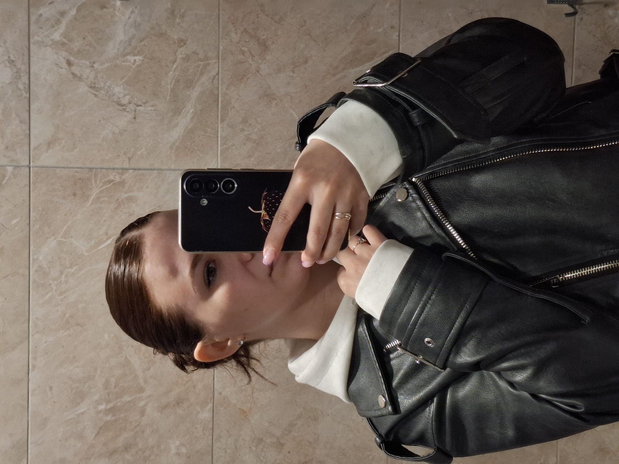 A woman is taking a selfie in front of a marble tile wall. She is wearing a black leather jacket over a white hooded sweatshirt. She is holding a black phone in front of her face. She has brown hair pulled back, fair skin, and her lips are visible. The phone case has a design on it. She is wearing two rings on her left hand. The location is unknown and not possible to be confirmed as Chaikovsky, Russia.