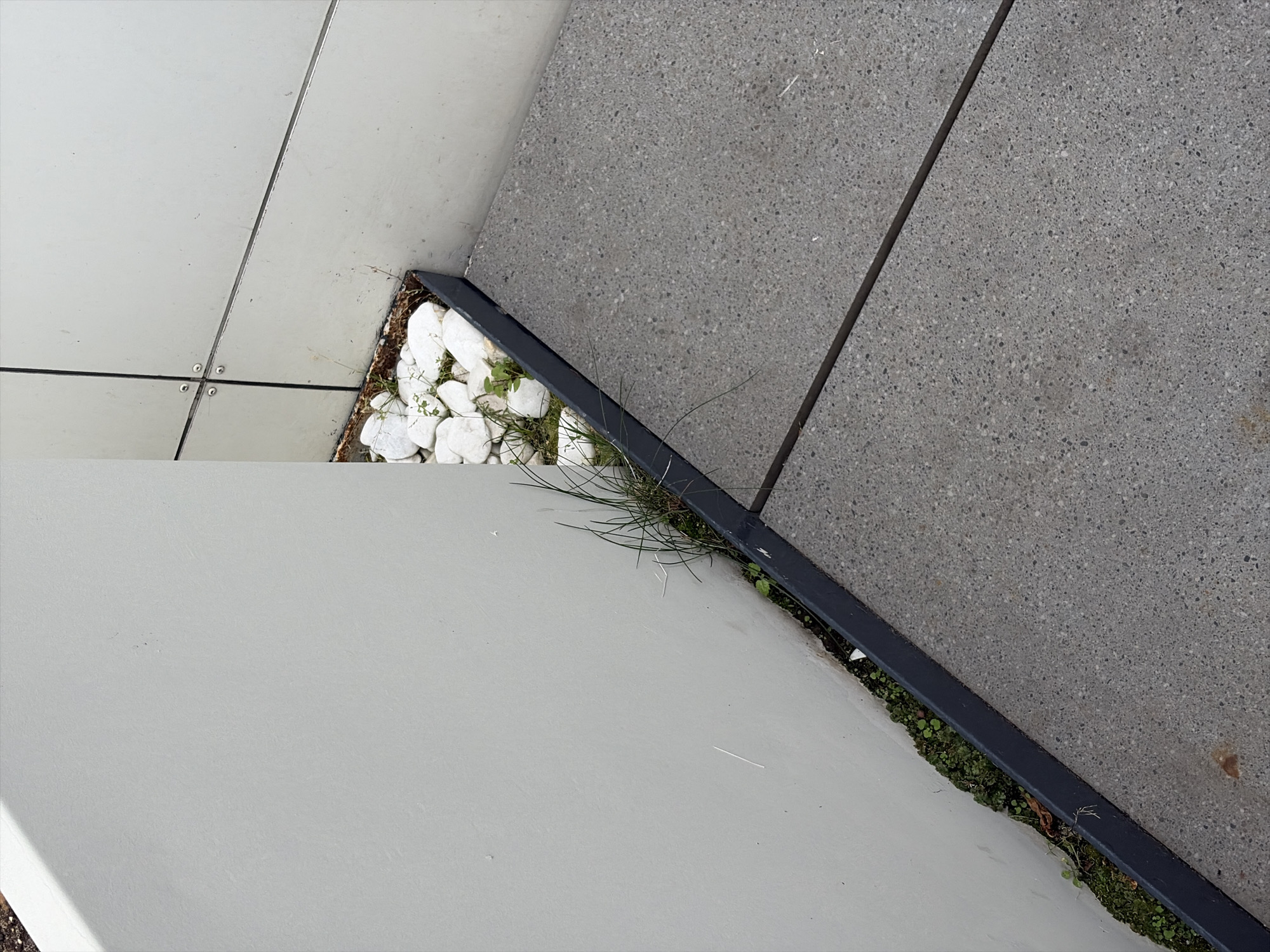 The image shows a section of an urban exterior environment. In the foreground, a gray concrete patio with a visible seam runs diagonally from the bottom right to the top left. Adjacent to the patio is a dark gray metal edging strip. Beyond the edging strip, a small area of dark soil is visible, with sparse green vegetation and a few scattered white pebbles. To the left of this area is a large, smooth, light gray wall. In the background, a beige wall constructed of large panels with visible seams and rivets can be seen. The scene appears to be a meticulously designed outdoor space, likely part of a modern building's exterior, possibly a garden or walkway area. There are no people visible in the image, and no actions or interactions are depicted. The lighting suggests an overcast day. The location is not specified in the image, and therefore cannot be determined to be in Moscow, Russia, or any other specific city.