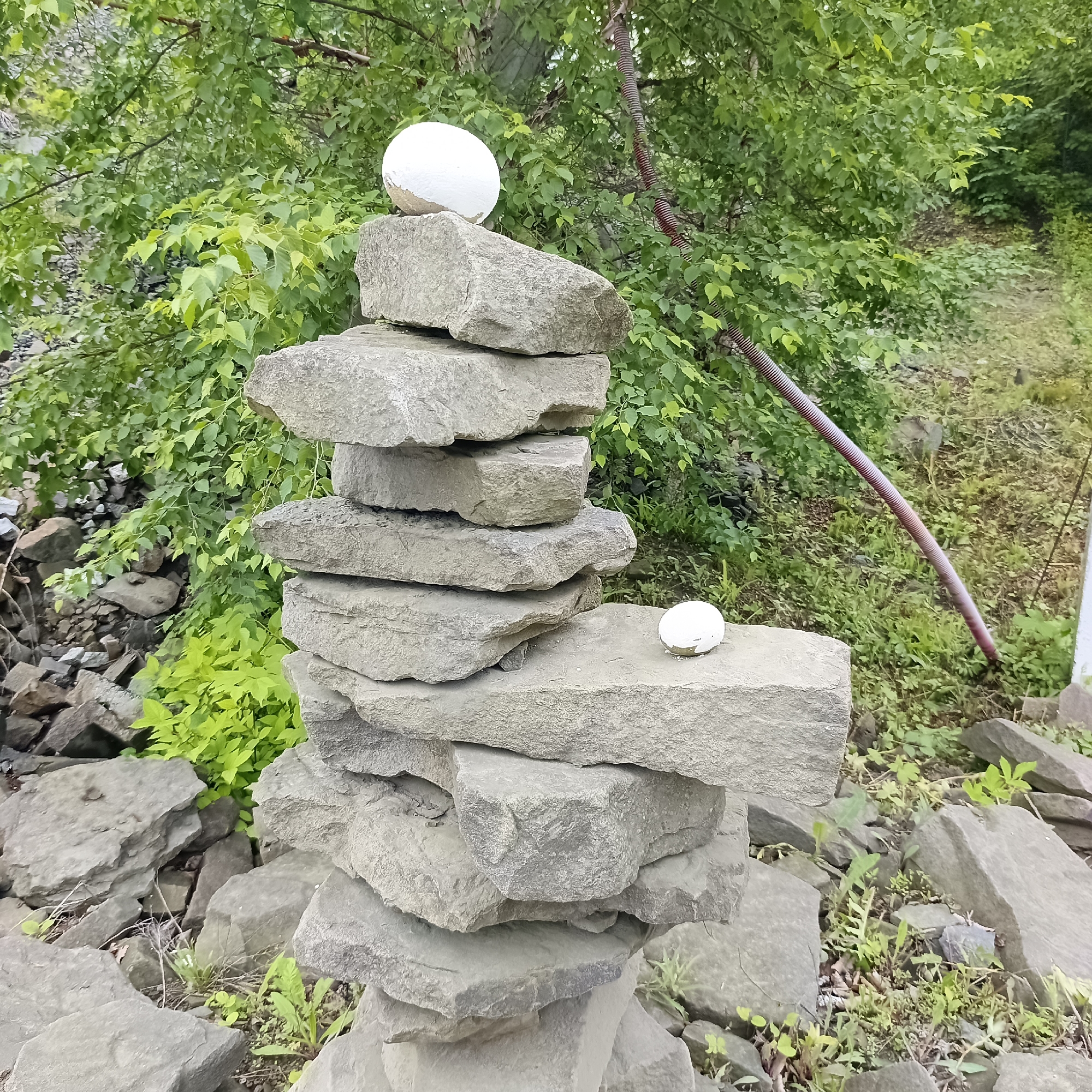A cairn made of flat, gray stones is stacked outdoors. Two white, spherical objects, possibly painted rocks or decorative orbs, are placed on the cairn. One is on the very top stone, and another is on a stone about halfway down. The cairn is situated among other scattered rocks and lush green foliage. In the background, a ribbed, reddish-brown pole is visible, leaning at an angle. The overall scene suggests a natural or semi-natural outdoor setting, possibly a park or a rocky landscape. The image does not contain any specific visual cues to determine its location within the city of Vladivostok, Russia.