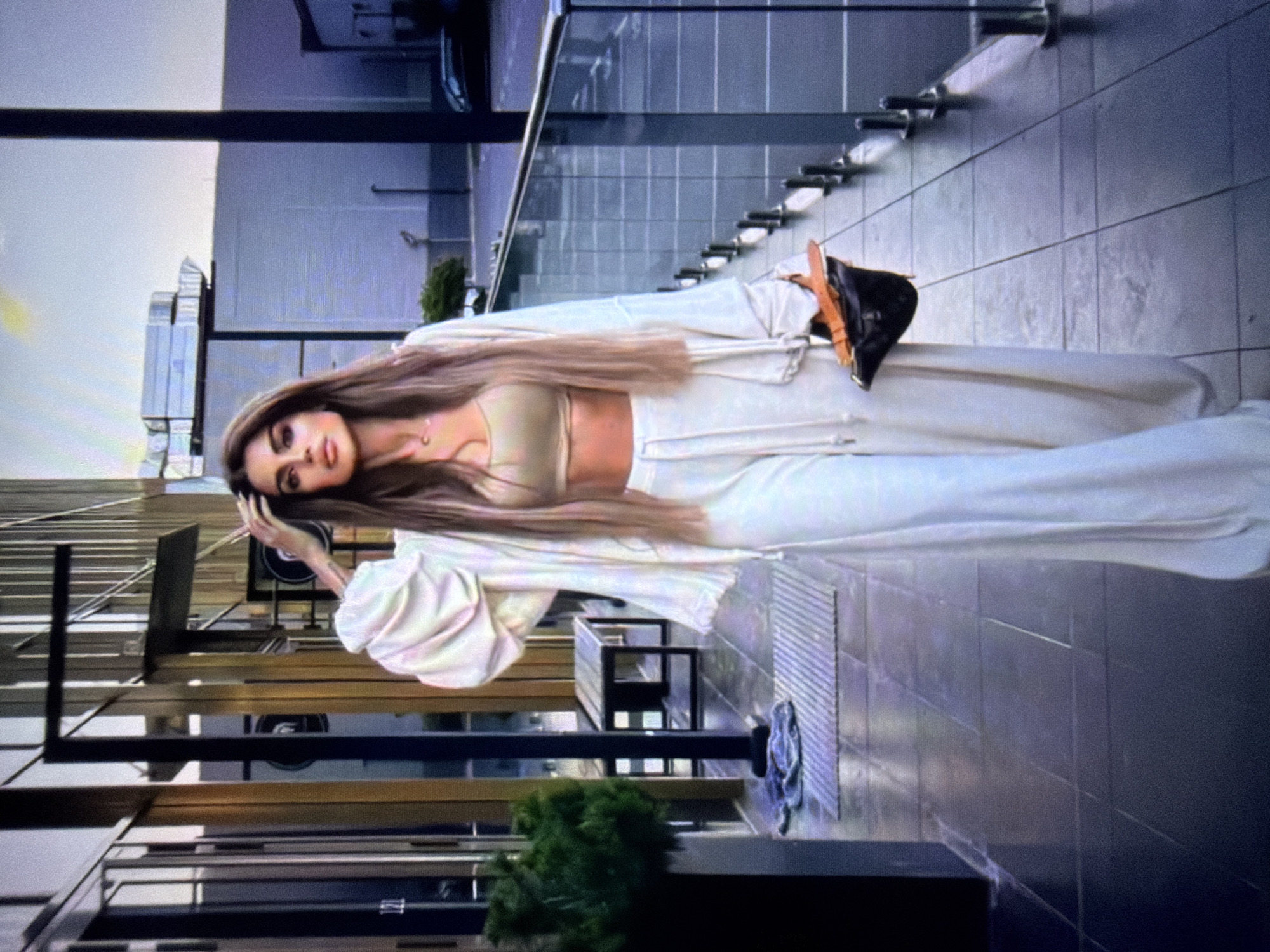 This media file shows a young woman posing outdoors in a modern setting. She has long, light brown hair, dark eyes, and is wearing a light-colored outfit consisting of a crop top and matching pants, along with a flowing jacket or robe. Her right hand is near her head, and she is looking towards the camera with a serious expression.
The scene appears to be a rooftop or elevated walkway with a tiled floor and a glass railing. In the background, there is a building with a facade of horizontally lined panels. Steps can be seen ascending on the right side of the frame. A small black modern-looking chair or table is also visible. The lighting suggests it is daytime, possibly late afternoon or early evening, with some natural light casting shadows. The overall atmosphere is chic and urban. There is no visible text in the image.