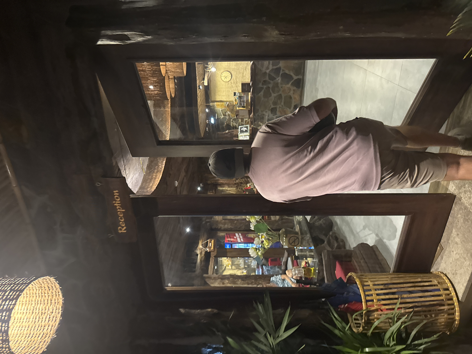 The image captures a person wearing a pink t-shirt and khaki shorts, with a dark baseball cap, walking towards a reception area. The reception area, visible through a large glass door and window, is characterized by its rustic, natural theme with stone walls and wooden elements. A wooden sign above the door reads "Reception" in an elegant font. Inside, a counter is manned by a person, and a clock hangs on the wall. The lighting is warm and inviting, with woven lampshades adding to the ambiance. In the foreground, to the left of the entrance, a bamboo basket holds several umbrellas, and lush green plants add a touch of nature. The overall impression is that of a tranquil and welcoming establishment, possibly a hotel or resort, in Nam Hoa Lư Ward, Vietnam.
