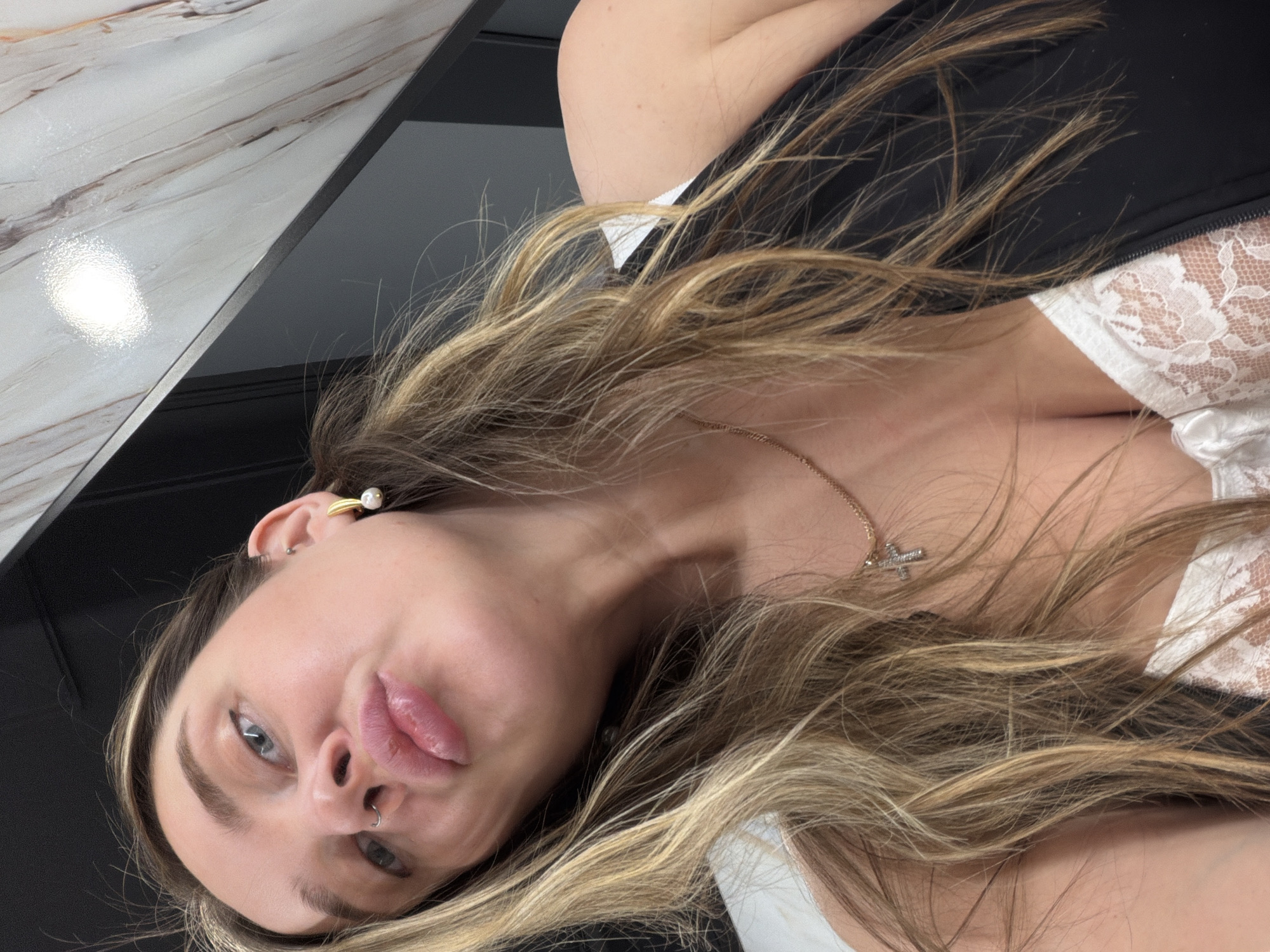 This close-up image, set in Moscow, Russia, features a young woman looking directly upwards at the camera. Her head is tilted, and she has a slightly puckered or pouty expression. She possesses long, wavy hair with blonde and light brown highlights, which flows around her face and across her chest.
Notable details on the woman include a small septum piercing, and on her visible ear, a gold-toned ear cuff or decorative earring featuring a pearl, along with a small stud. She wears a delicate gold-toned chain necklace adorned with a small, sparkling cross pendant. Part of a white lace bra or top is visible on her chest, worn beneath a dark, possibly black, garment.
The background consists of a glossy, light-colored surface with a marble-like pattern (white with reddish-brown veins) on the upper left, reflecting a bright light. Adjacent to this is a dark, smooth, likely black or dark grey surface. These surfaces suggest a contemporary indoor setting, possibly a countertop or table. The image captures a posed moment, conveying a sense of playfulness and confidence. No visible text is present in the media file.
