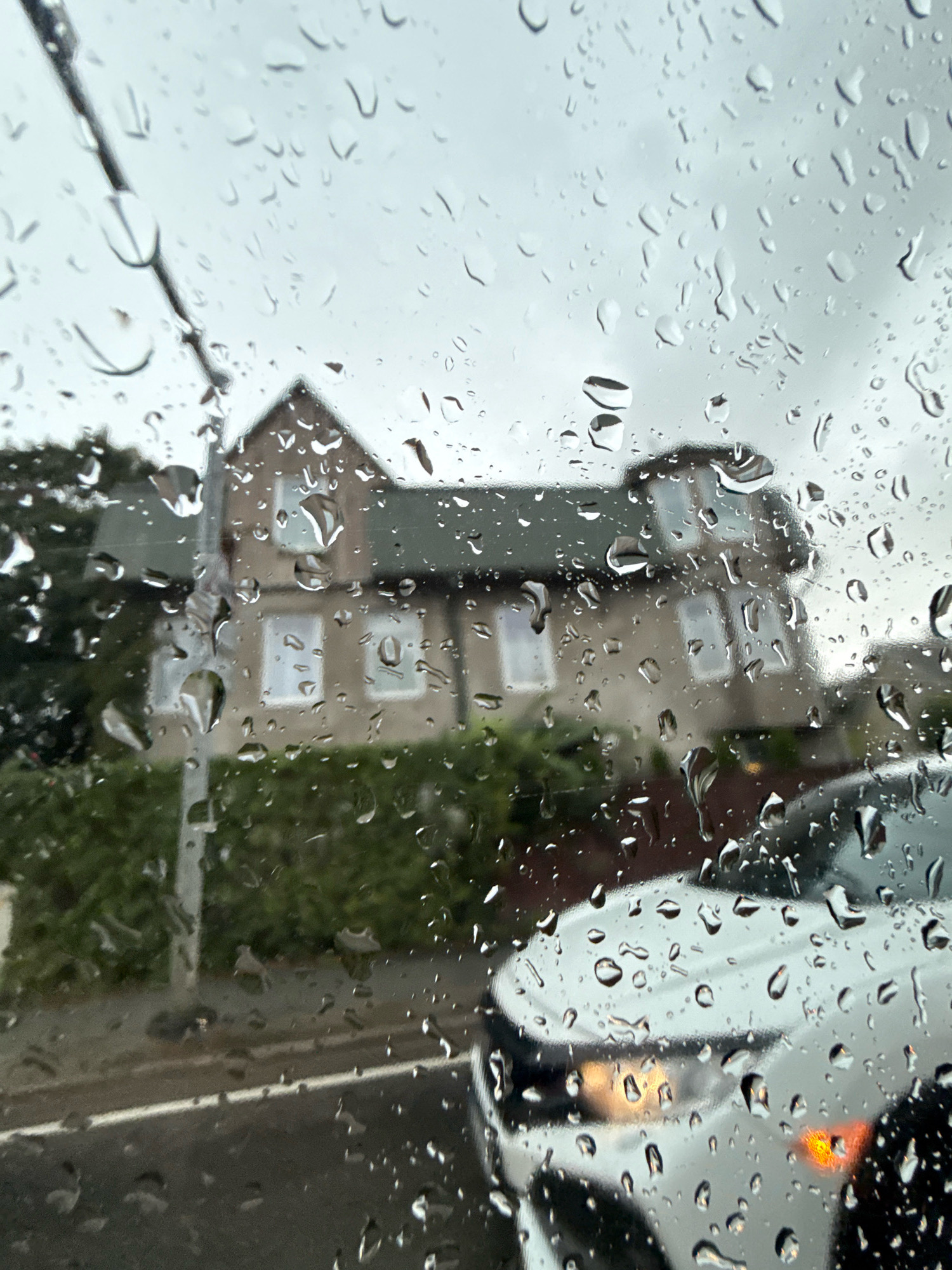 The image is a view through a rain-streaked window of a house and a white car on a cloudy day. The house is a two-story building with a dark green roof and beige walls, featuring several windows. A hedge runs along the front of the property. A white car is visible in the foreground, with its headlights on. The overall atmosphere is somber due to the overcast sky and rain. The image does not provide any specific clues about the location in Tallinn, Estonia.