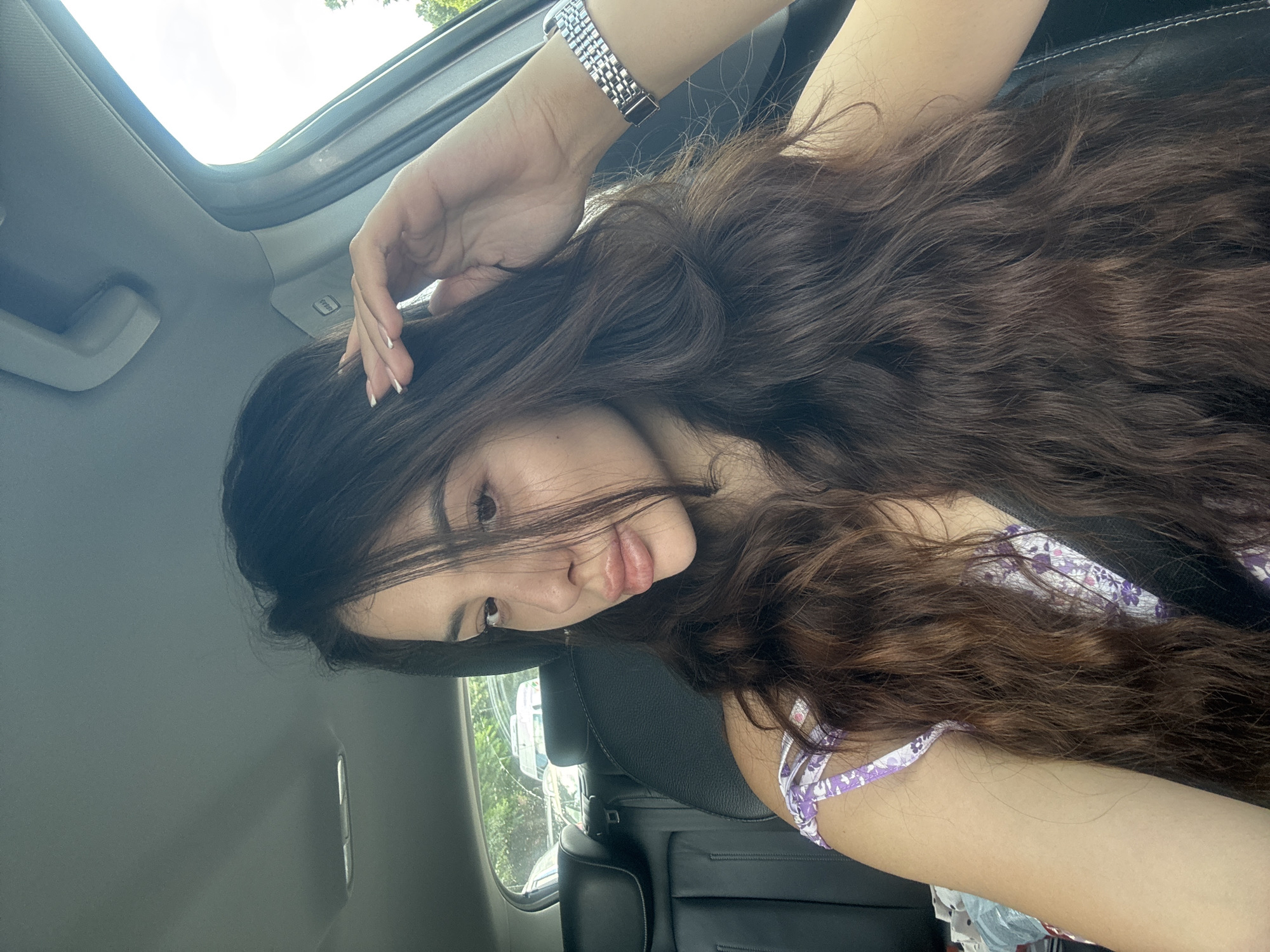 The image depicts a woman inside a car. She has long, wavy brown hair and is wearing a watch. Her hand is touching her hair. The car appears to have a sunroof. The location is not apparent from the image, and there is no indication this image was taken in the city of Plovdiv, Bulgaria.