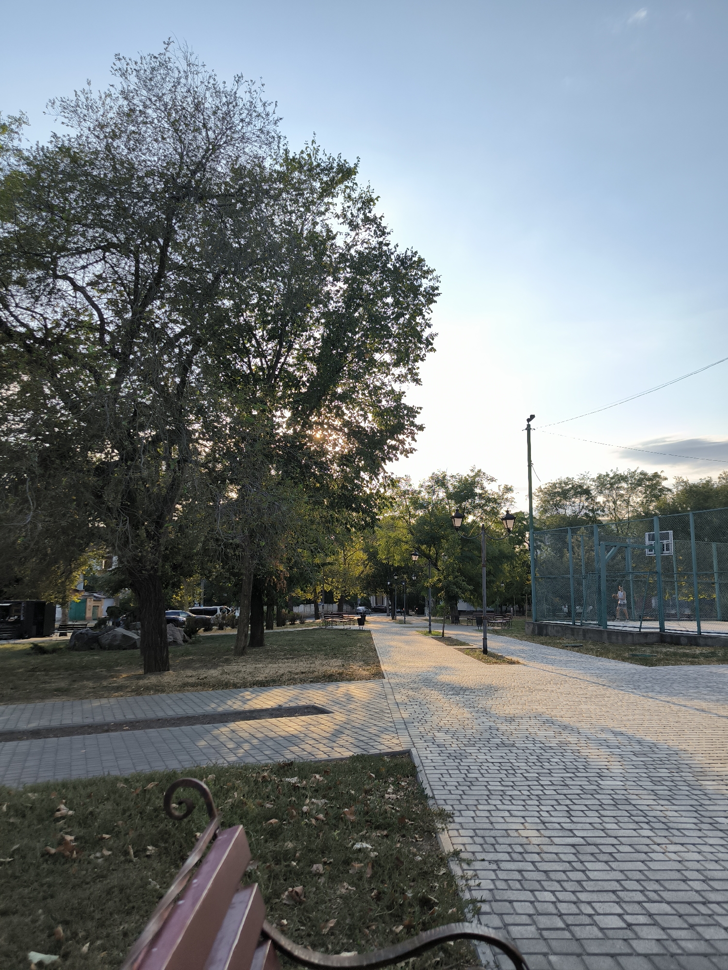 The image depicts a park pathway lined with trees on the left and a sports court on the right. The pathway is paved with light-colored cobblestones, and dappled sunlight casts shadows across it. Several streetlights with ornate designs are visible along the path. The sports court, enclosed by a green metal fence, appears to be a basketball court, with a lone individual visible on it. Trees with lush green foliage flank the pathway, and some dry leaves are scattered on the grassy areas. In the foreground, a portion of a park bench with a metal frame and reddish-brown slats is visible. The sky is clear and blue, with a hint of clouds towards the horizon. The scene suggests a peaceful, late afternoon or early evening setting in a public park. The presence of the park benches, pathway, and trees, combined with the sports court, indicates a recreational area within a city. The overall ambiance is serene and inviting. This scene is a typical view of a public park in Mykolaiv, Ukraine, designed for recreation and relaxation.