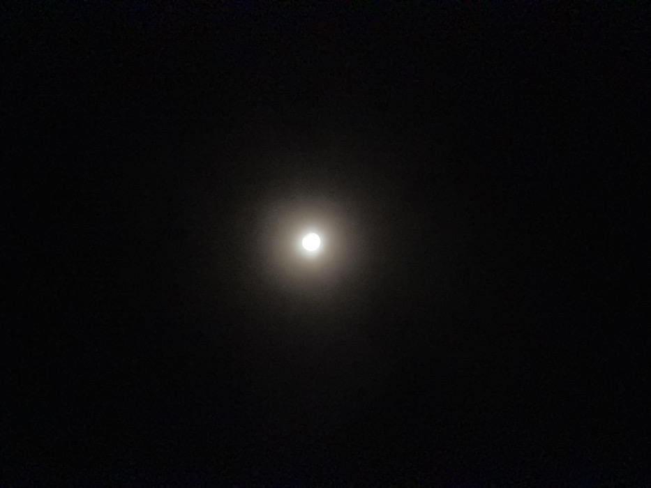 The image depicts a single, bright, spherical celestial body, identified as the moon, centered against a uniformly dark, featureless night sky. A soft, diffuse halo of light surrounds the central luminous object. No other celestial bodies, clouds, terrestrial features, structures, individuals, or objects are discernible within the frame. The scene is located in Kemenuh, Indonesia. No actions or interactions are visible.