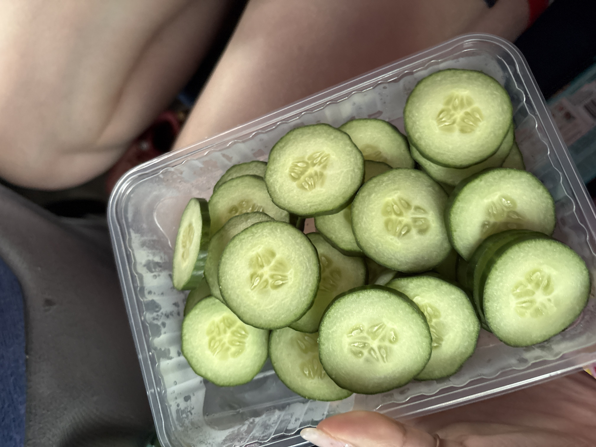 The image shows a clear plastic container filled with sliced cucumbers. The container is being held by a person, whose fingers are visible. The background includes clothing and possibly a leg. The location is unknown, and there is no verifiable information to suggest it is in Yanychi, Russia.