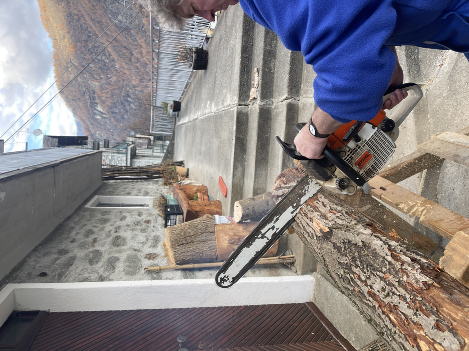 The image captures an outdoor scene in Dosso (Ciappanico), Italy, depicting a man cutting wood with a chainsaw. A person, seen from the chest up, wearing a blue fleece jacket, is actively operating a Stihl chainsaw. His hands, one gloved and the other bare, wearing a black watch, firmly grip the saw as he cuts a large, irregularly shaped log resting on a wooden stand. Sawdust is visible around the cutting area.
The scene is set on a concrete or paved outdoor terrace, with a stone wall and concrete steps ascending in the background. A metal railing is visible at the top of the steps. Below the cutting area, a stone building facade features a window with a white frame. Further in the background, several traditional buildings with grey roofs and stone or concrete walls are nestled into the steep terrain, characteristic of a mountain village. The distant mountain is covered in dense, autumnal foliage, displaying shades of brown and orange under a partly cloudy sky. Other cut logs are stacked nearby, suggesting ongoing wood processing. The activity takes place during daytime. The only visible text is "Stihl" on the chainsaw.