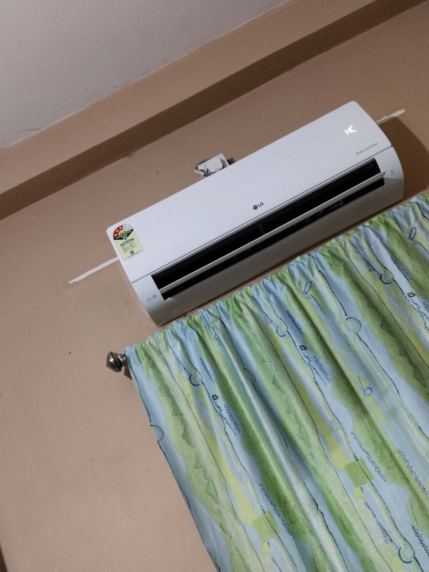 An indoor shot captures an air conditioning unit mounted on a light brown wall, partially obscured by a curtain with a green and blue abstract pattern. The air conditioner is white and features the LG logo. The display on the unit shows "HC" and "DUAL Inverter". A yellow sticker with star ratings is also visible on the unit. The room appears to be a residential setting in Rajpur Sonarpur, India.