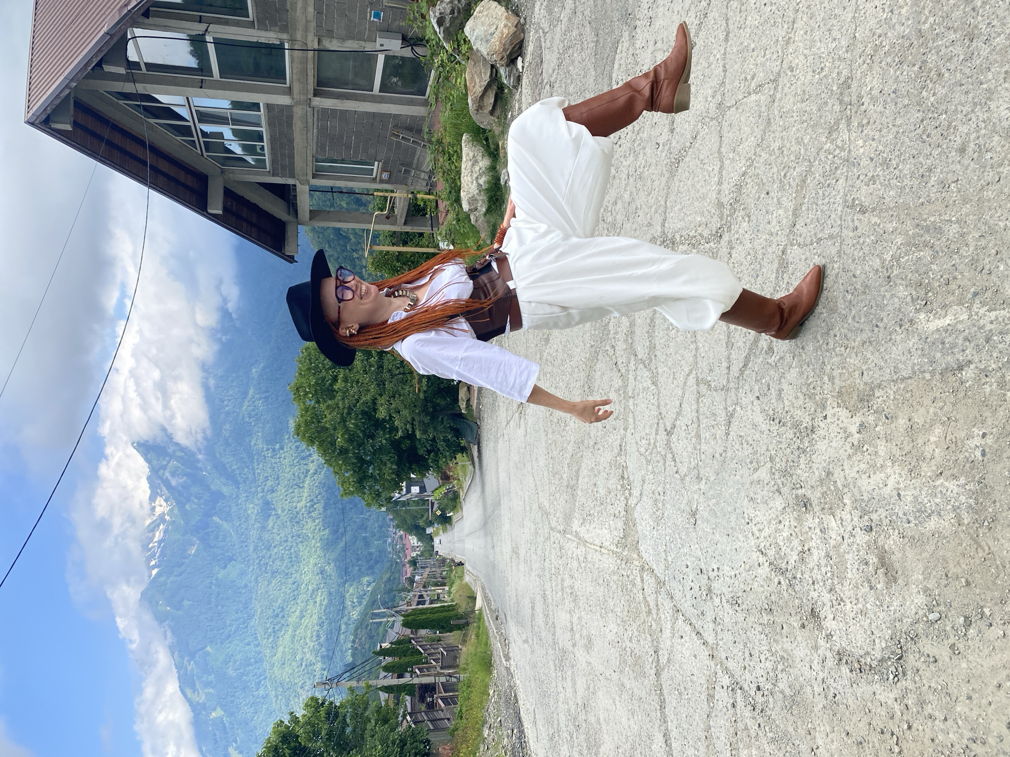 A woman with long, braided orange hair, wearing glasses, a black hat, a white shirt, white pants, a brown corset, and brown boots is posing for a photo. She appears to be floating in the air, with her legs extended and her arms outstretched. She is smiling and looking at the camera.
The scene is outdoors, on a paved road that slopes downhill. In the background, there is a building with a grey concrete exterior and a brown roof, a lush green mountainside with some snow-capped peaks in the distance, and a village nestled in the valley. The sky is blue with some white clouds. The lighting suggests it is daytime, and the weather appears to be pleasant.
The image evokes a sense of freedom and joy, with the woman's playful pose and the beautiful natural scenery. The location, Krasnaya Polyana, Russia, is known for its mountainous landscape and ski resorts, which aligns with the visual cues in the image.