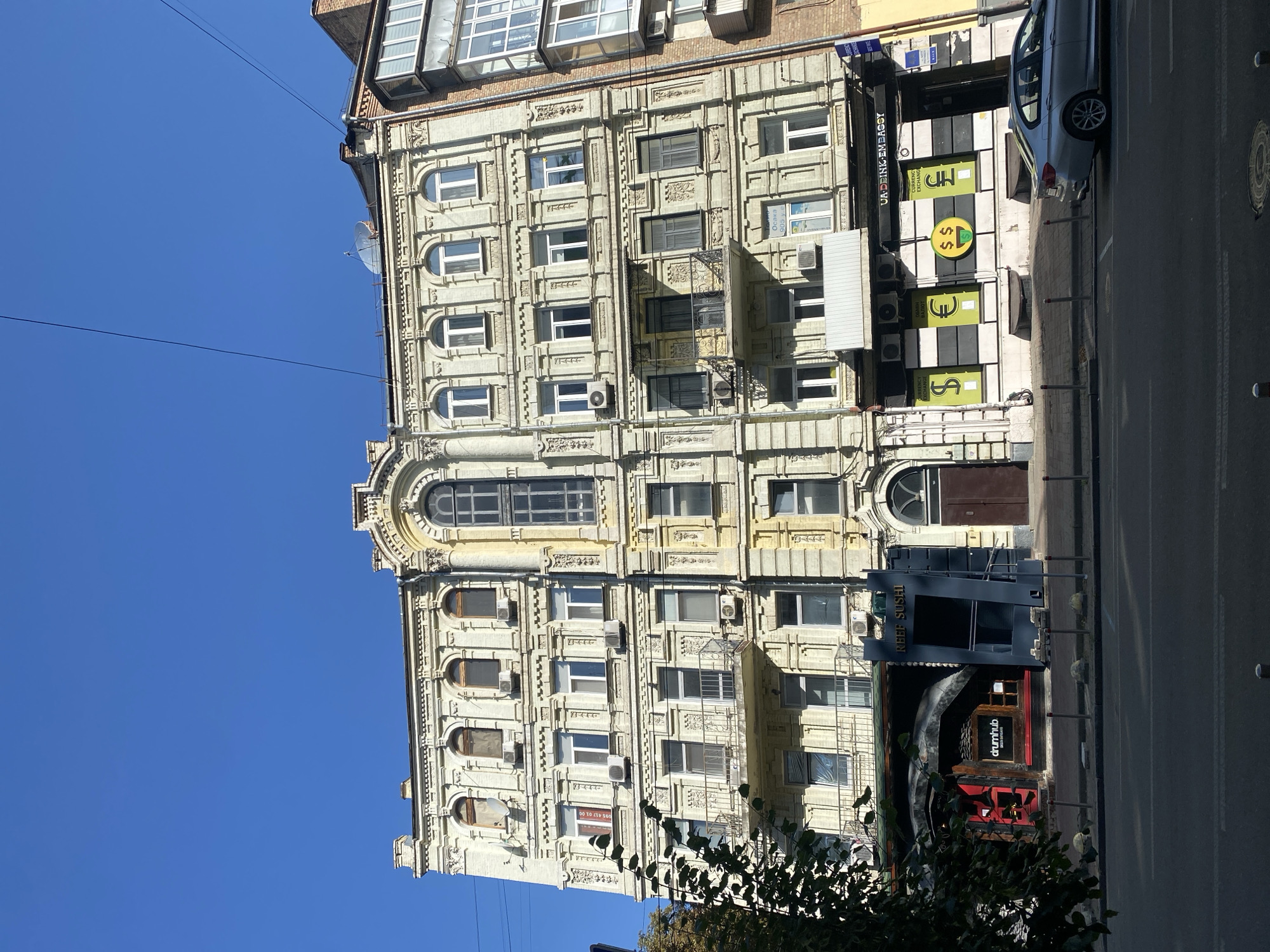 This is an outdoor, eye-level, full shot of a light yellow, four-story building with an ornate facade and a clear blue sky above. The building is located in an urban setting, with a street in the foreground. The building has many windows, some with arched tops, and balconies with decorative iron railings. Several air conditioning units are visible on the exterior. The ground floor of the building houses several businesses, including a restaurant with a red entrance and a sign that reads "drumhub," and a currency exchange with signs displaying dollar, euro, and pound symbols. A silver car is parked on the street in front of the currency exchange. Trees with green foliage partially obscure the left side of the building. The overall impression is one of a well-maintained, historic urban building.