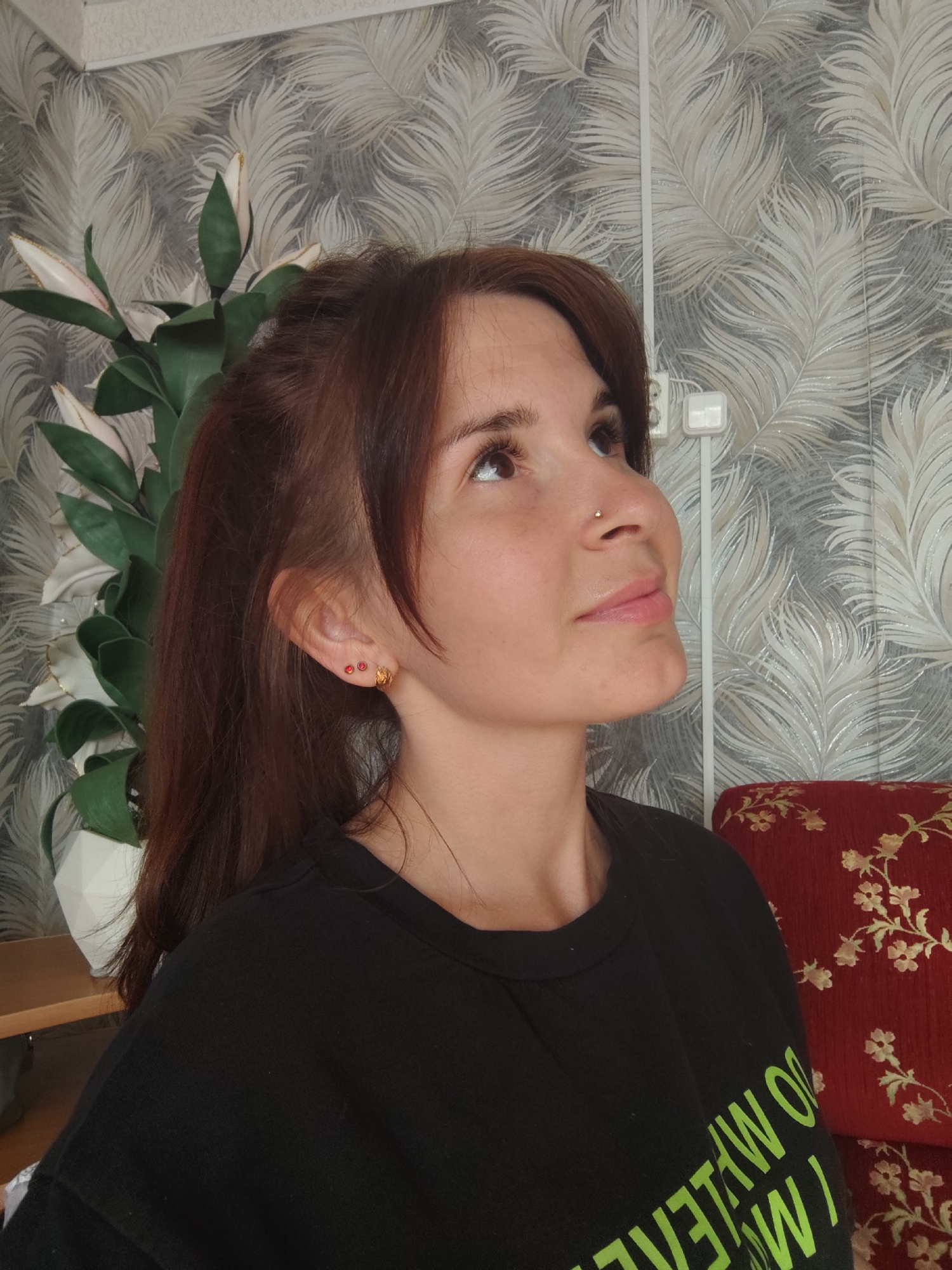A woman with brown hair and a nose piercing looks upward. She wears a black shirt. A plant with white flowers is to her left. The wall behind her features a feather pattern. A red cushion with a floral pattern is on the right. The location is possibly Альняш, Russia.