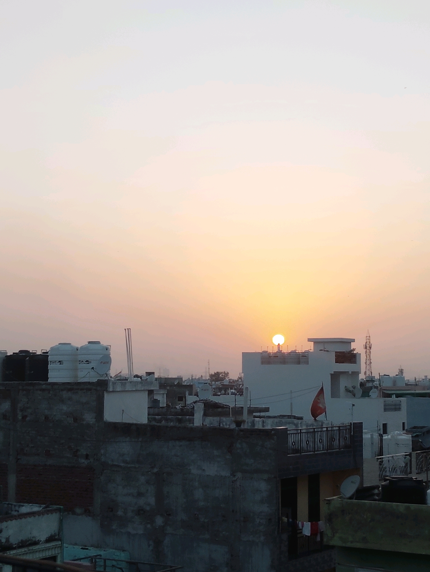 This image depicts a rooftop view of a cityscape at sunset in Delhi, India. The sky is dominated by warm hues of orange and yellow, indicating the sun is low on the horizon. Several buildings with flat roofs are visible, typical of urban architecture in Delhi. On some of these roofs, large white water tanks, common for storing municipal water, are present. In the mid-ground, a building with a white facade stands out, and atop it, a red flag with an emblem is prominently displayed. Antennas and satellite dishes are also visible on various rooftops, suggesting residential or commercial activity. The foreground shows the weathered texture of older buildings, with visible brickwork and concrete. The overall scene captures a typical urban environment in Delhi during twilight, with an emphasis on the residential structures and the atmospheric conditions of the setting sun. There are no visible people in the image.