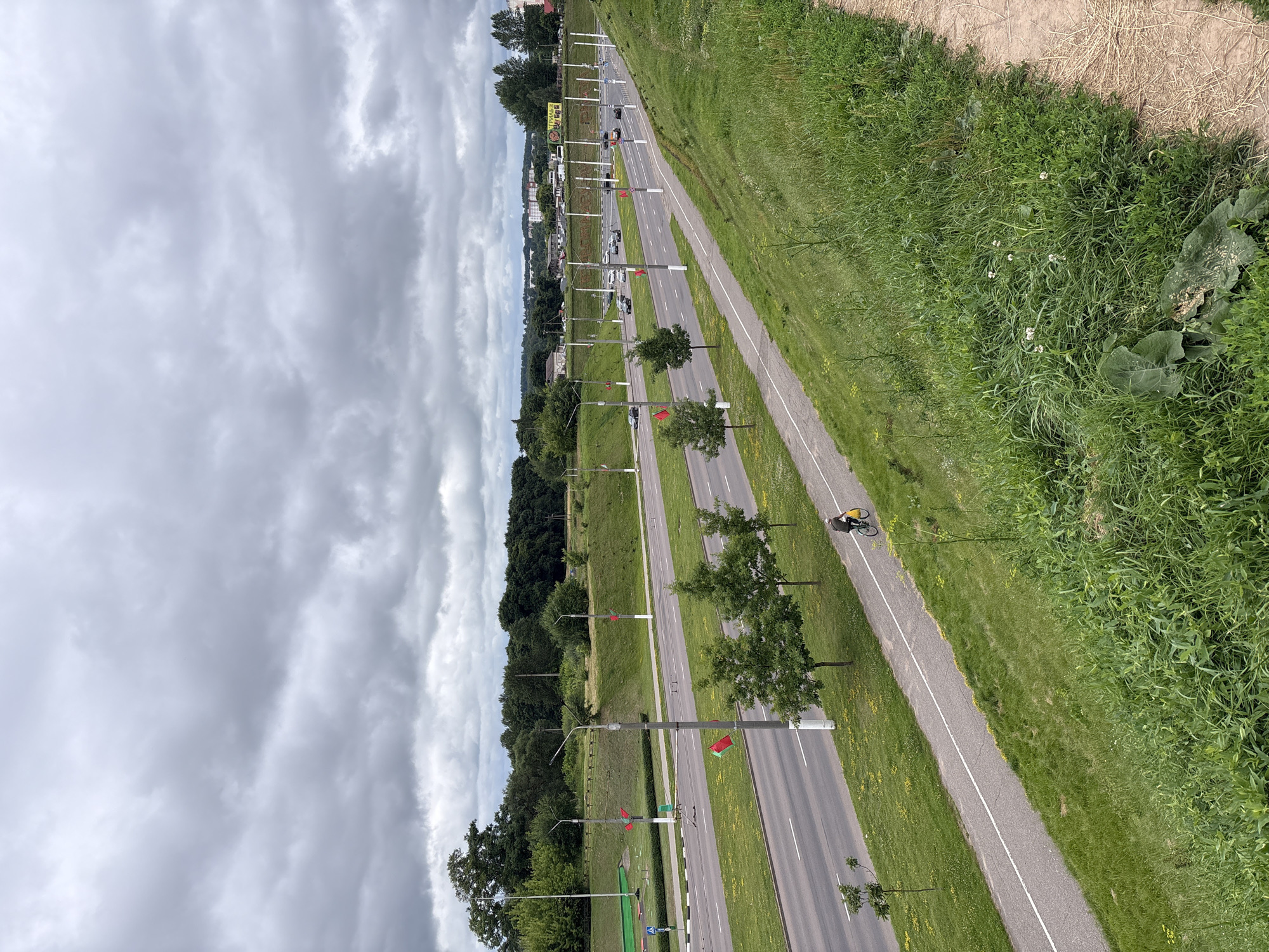The image depicts a wide, multi-lane road in Vitsebsk, Belarus, on a cloudy day. A bicycle path runs parallel to the road on the right side, separated by a grassy embankment. A lone cyclist with a yellow bag is riding on the path. Several trees are planted along the median and the grassy areas. Red and green flags are visible on poles lining the road. In the distance, buildings and more trees are visible. The overall scene suggests an urban or suburban setting with a focus on transportation infrastructure.